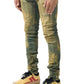 A person wearing distressed SERENEDE KALAMATA JEANS OLIVE BROWN with a dirty washed effect in green and yellow, paired with yellow sneakers featuring red stripes, stands against a white background.