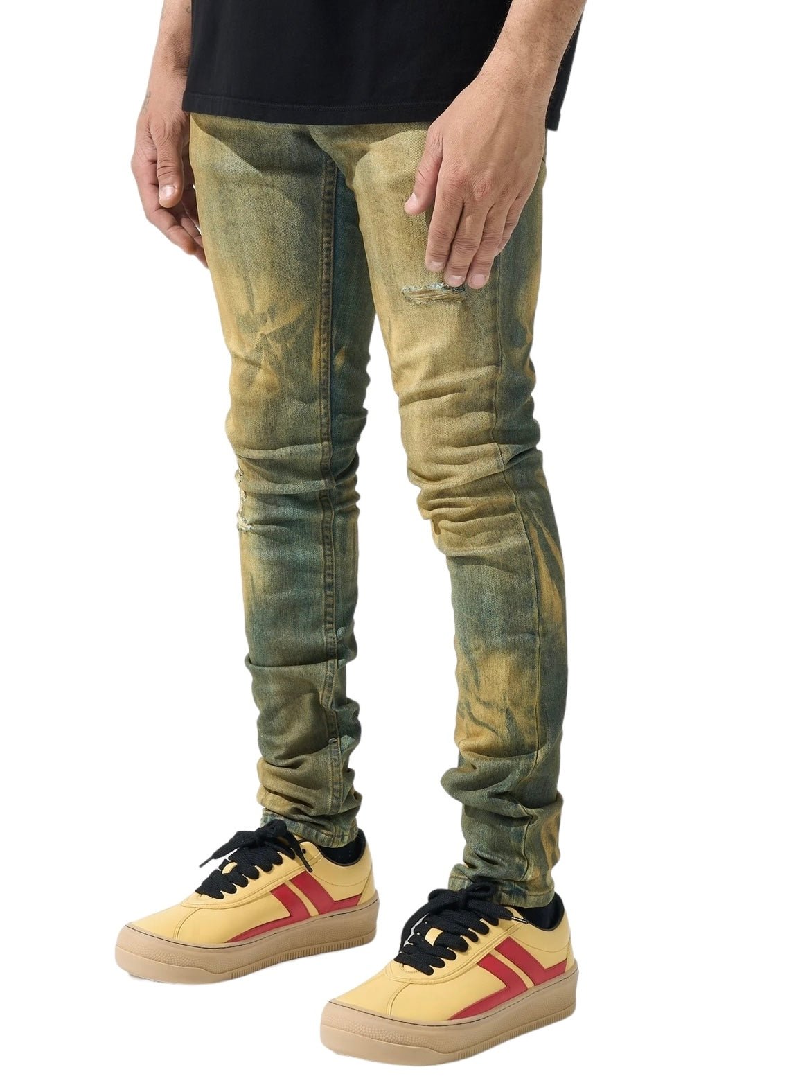 A person wearing distressed SERENEDE KALAMATA JEANS OLIVE BROWN with a dirty washed effect in green and yellow, paired with yellow sneakers featuring red stripes, stands against a white background.
