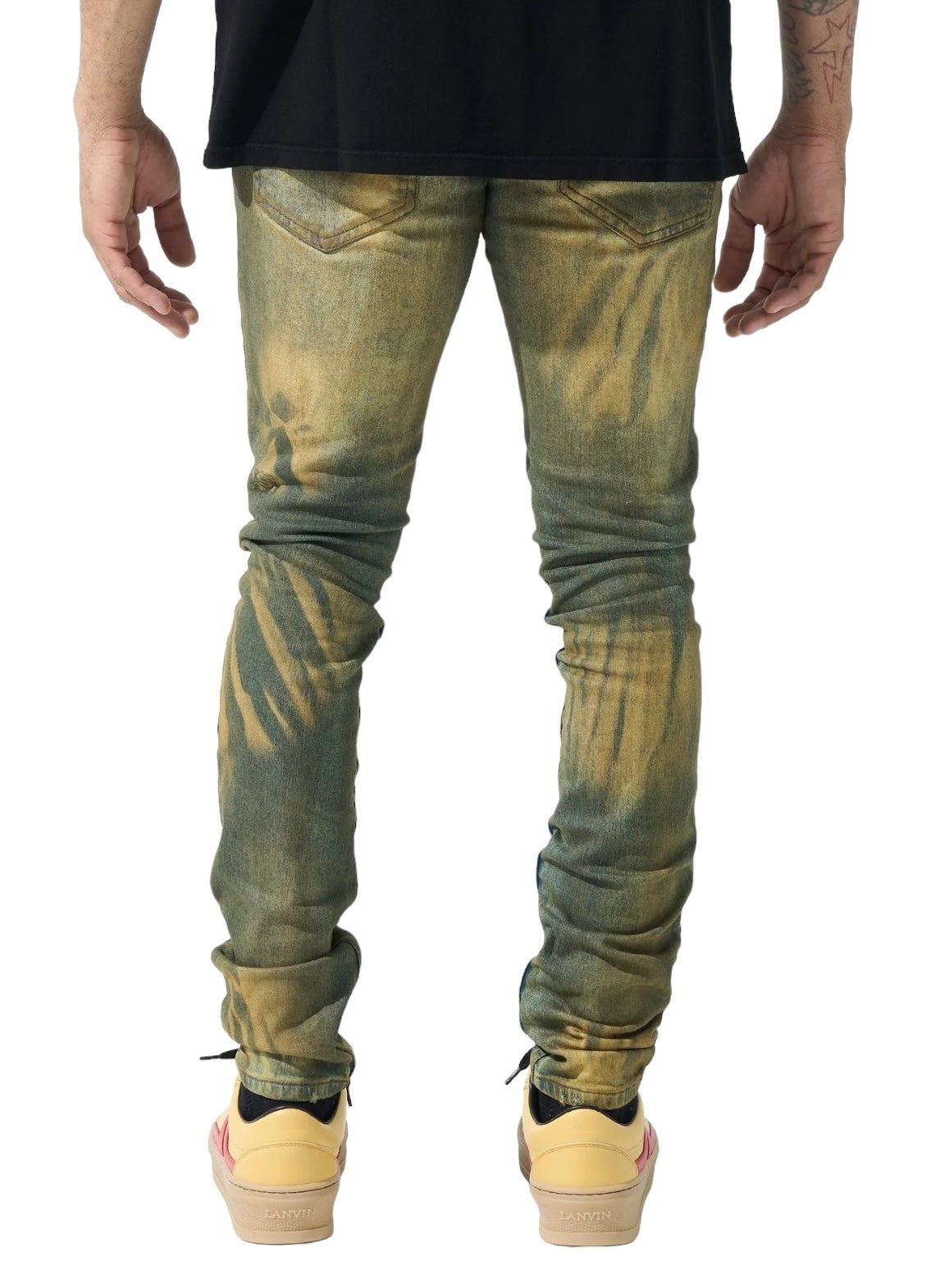 Back view of a person wearing SERENEDE KALAMATA JEANS OLIVE BROWN and beige sneakers, with tattoos visible on one arm. The person is also sporting a black top.