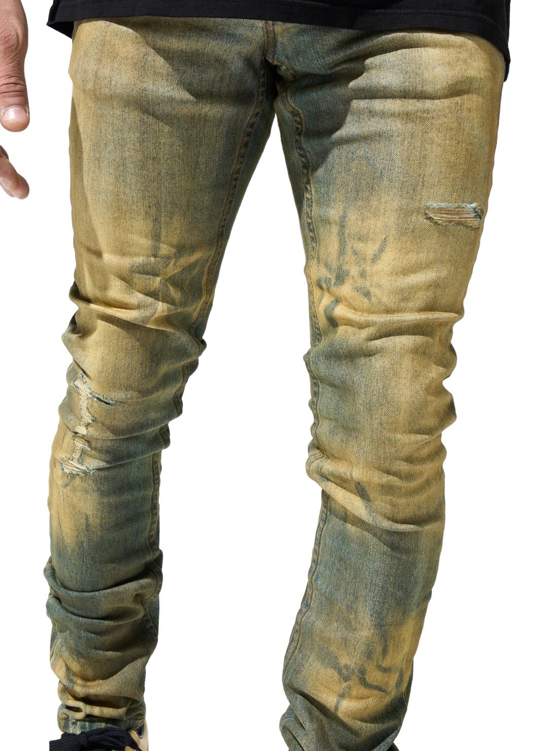 Person wearing distressed, skinny-fit SERENEDE KALAMATA JEANS OLIVE BROWN with a dirty washed effect and visible tears, paired with a black top, showing only the legs and a partial hand on the left side.