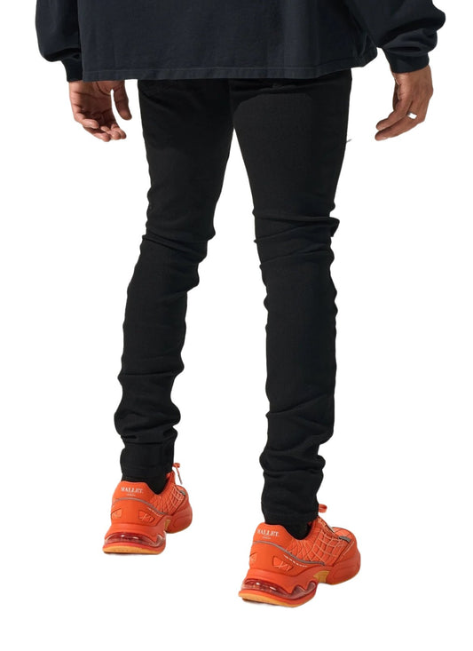 A person wearing SERENEDE MIDNIGHT BLACK JEANS and vibrant orange sneakers against a white background.