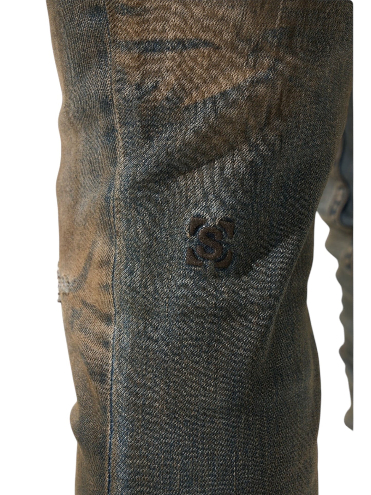 Close-up of SERENEDE Serenede Oud Jeans Oudwood, featuring distressed, dark-washed skinny fit with a black embroidered floral design on the thigh. Crafted from cotton, these jeans showcase textured brown and blue dye patterns.