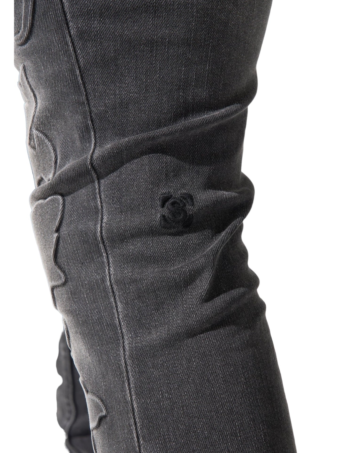 Close-up photo of a person wearing dark, distressed SERENEDE ROME JEANS BLACK with noticeable stitching details and a small embroidered patch featuring a vintage wash and skinny fit.