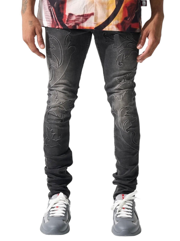 A person stands wearing black SERENEDE ROME JEANS with an intricate embroidered pattern and gray sneakers with white laces.