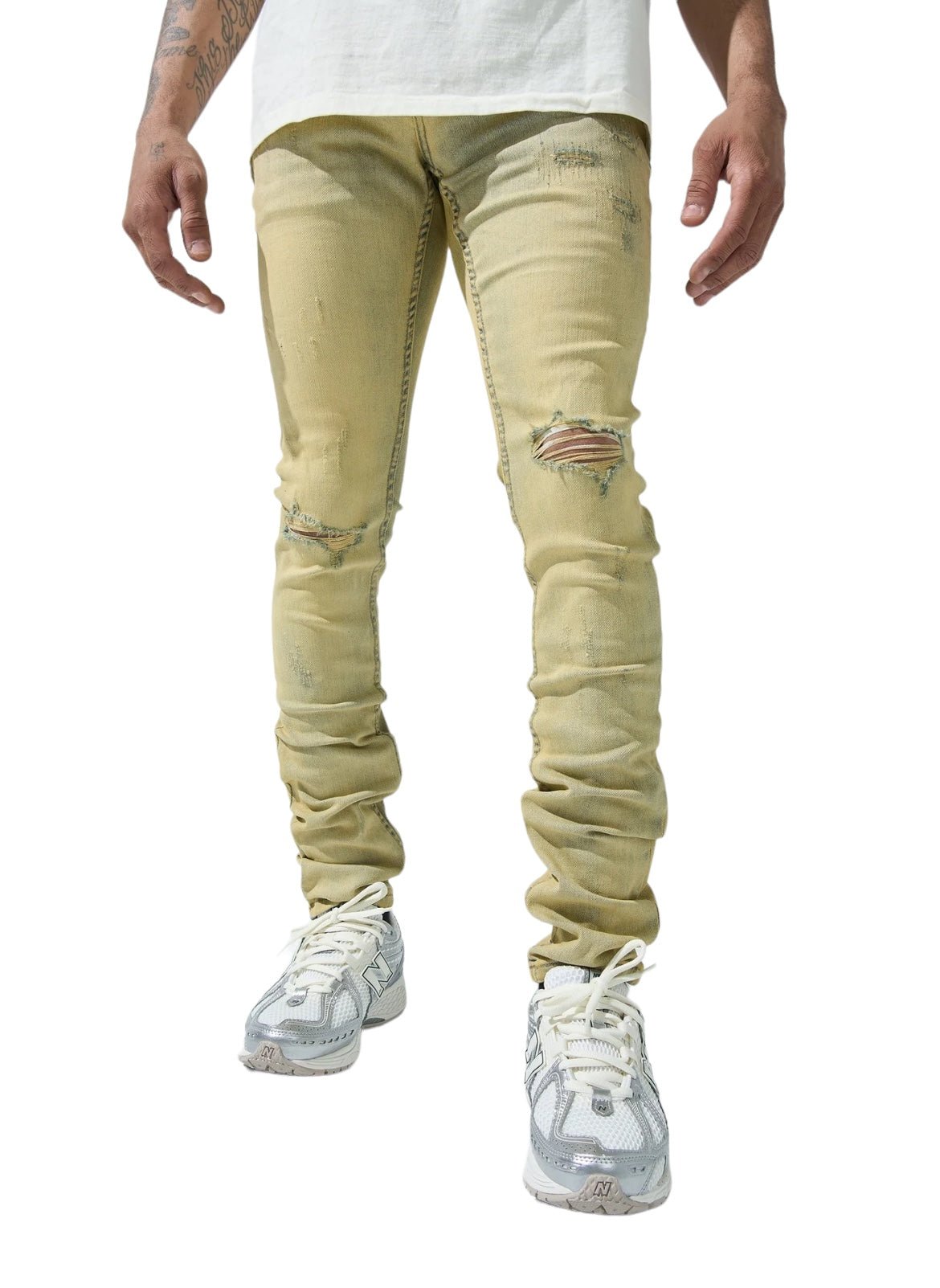 A person wearing SERENEDE SOL JEANS RISE, a pair of distressed, light beige men's jeans made of stretch cotton with rips on the knees, a white shirt, and white athletic shoes. The photo shows the lower half of the body.