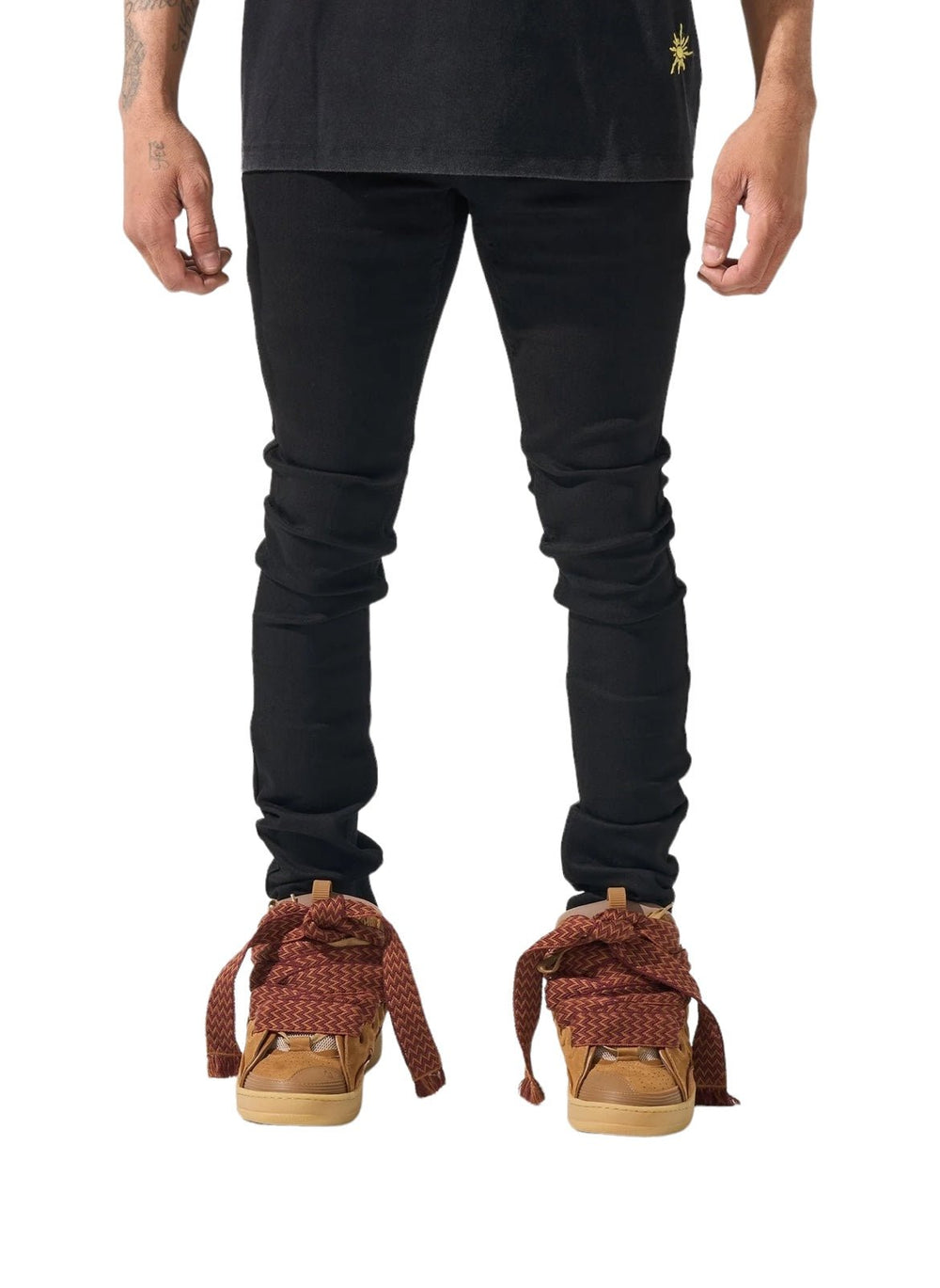 The man is dressed in black pants and a black shirt, complemented by brown shoes oddly tied with long, wide red laces. His hands hang casually at his sides, the sleek look of his SERENEDE VANTA 11 JEANS BLACK crafted from premium fabric adding a sophisticated touch.