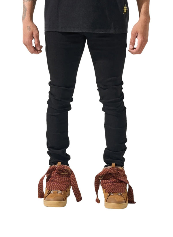 The man is dressed in black pants and a black shirt, complemented by brown shoes oddly tied with long, wide red laces. His hands hang casually at his sides, the sleek look of his SERENEDE VANTA 11 JEANS BLACK crafted from premium fabric adding a sophisticated touch.