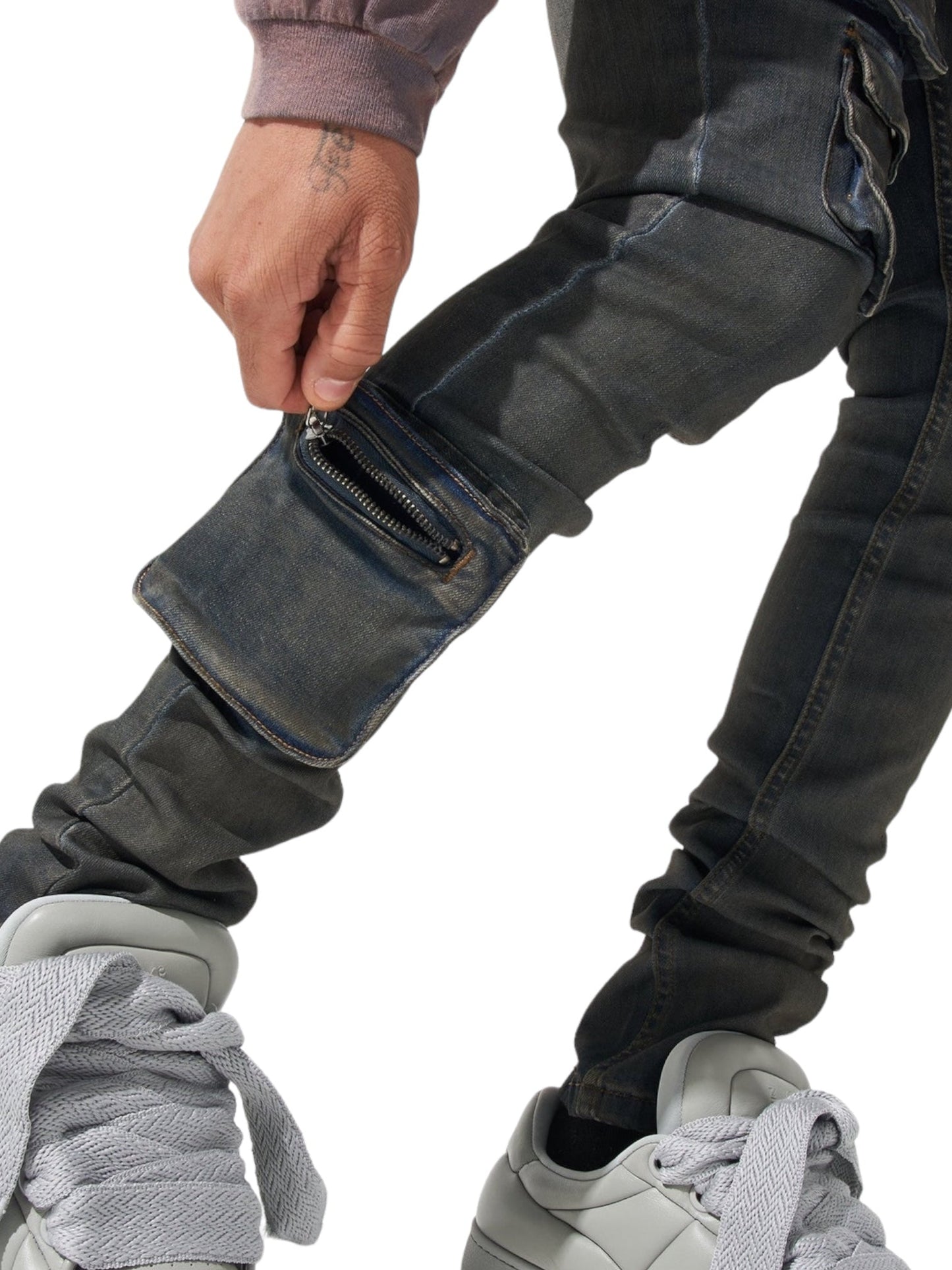 Close-up of a person wearing SERENEDE WAVE CARGO JEANS from the brand SERENEDE. The dark, slightly distressed jeans in the Atlantic dark blue shade feature a zipper pocket on the leg. The person pairs them with large, light gray sneakers with thick laces and is seen pulling at the jeans near the zipper.