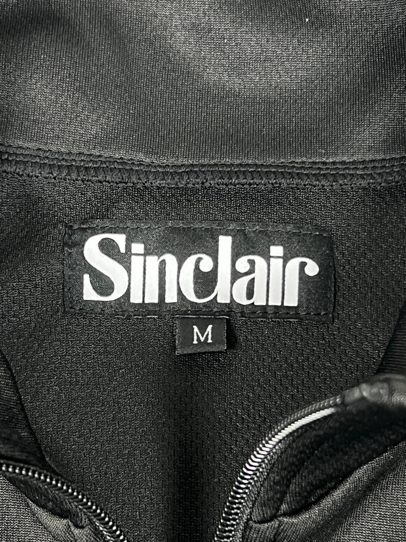 Close-up of a black SINCLAIR SMT190 Tech Logo Track Jacket featuring an embroidered "Sinclair" logo and a size "M" tag.