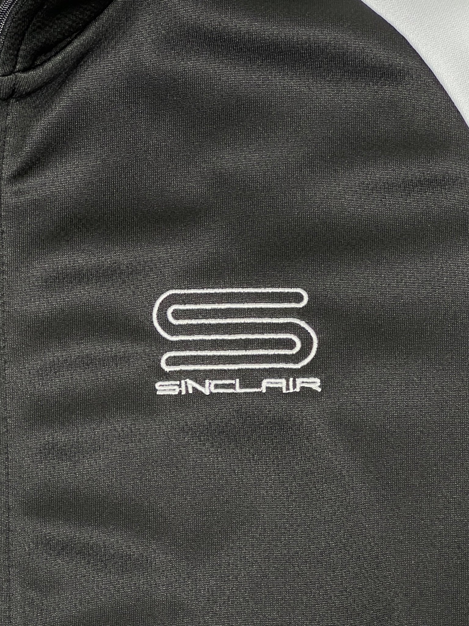 A close-up of the Sinclair SMT190 Tech Logo Track Jacket Black highlights the SINCLAIR logo: two stacked horizontal lines with "SINCLAIR" in white, expertly embroidered on the garment.