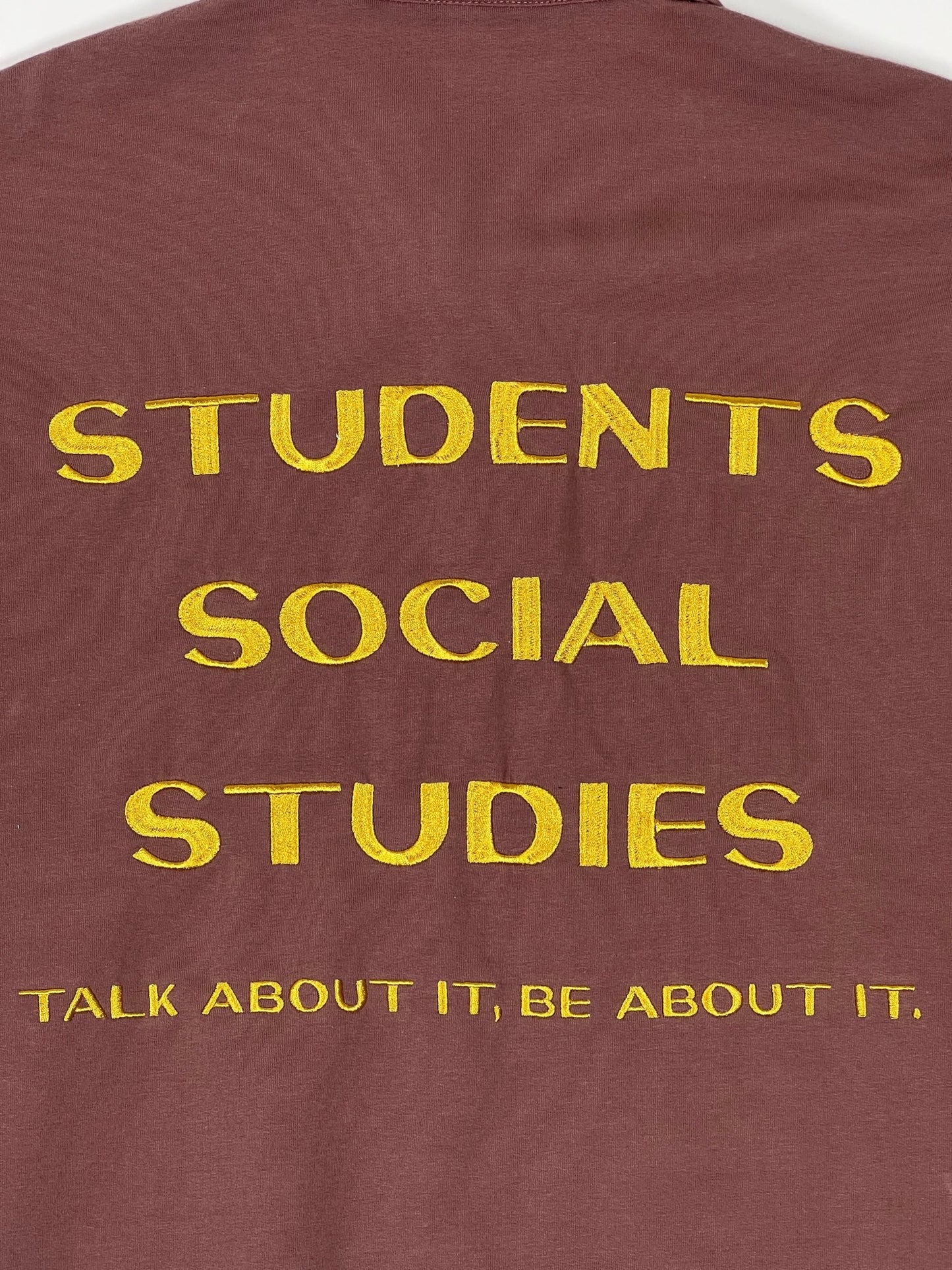 Brown Callum long-sleeve polo shirt by STUDENTS, made from soft French terry cotton and featuring gold screen-printed text: "SOCIAL STUDIES TALK ABOUT IT, BE ABOUT IT.