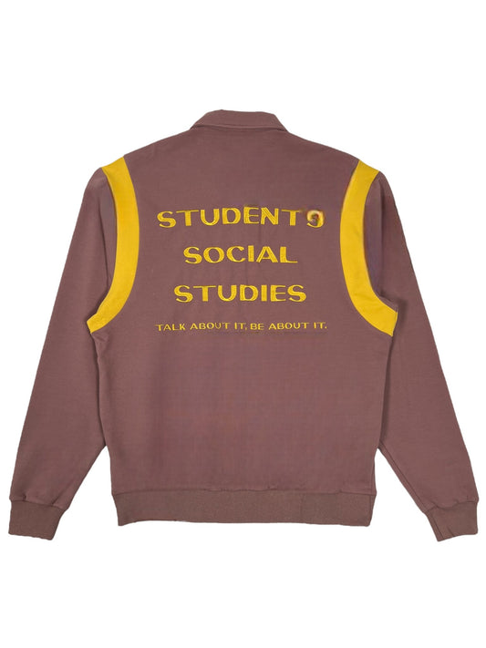 Introducing the Students Callum long-sleeve polo shirt in brown by STUDENTS. It features yellow stripes on the shoulders and is made from comfortable French terry cotton. A screen-printed design on the back reads "STUDENT9 SOCIAL STUDIES TALK ABOUT IT, BE ABOUT IT.