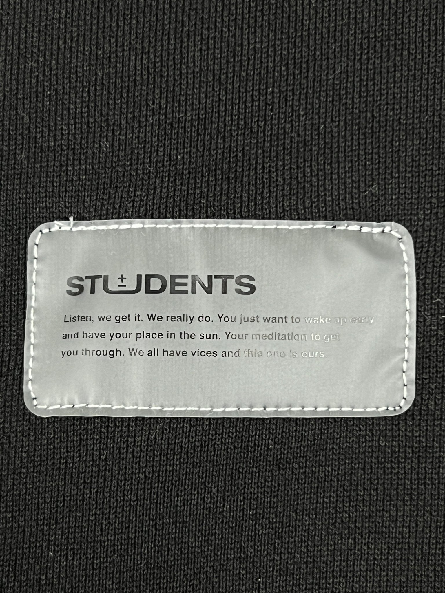Close-up of a fabric label on the "Students Estudiantes Pullover Hood Blk" by STUDENTS with a motivational message, sewn onto 100% cotton black hoodie material.