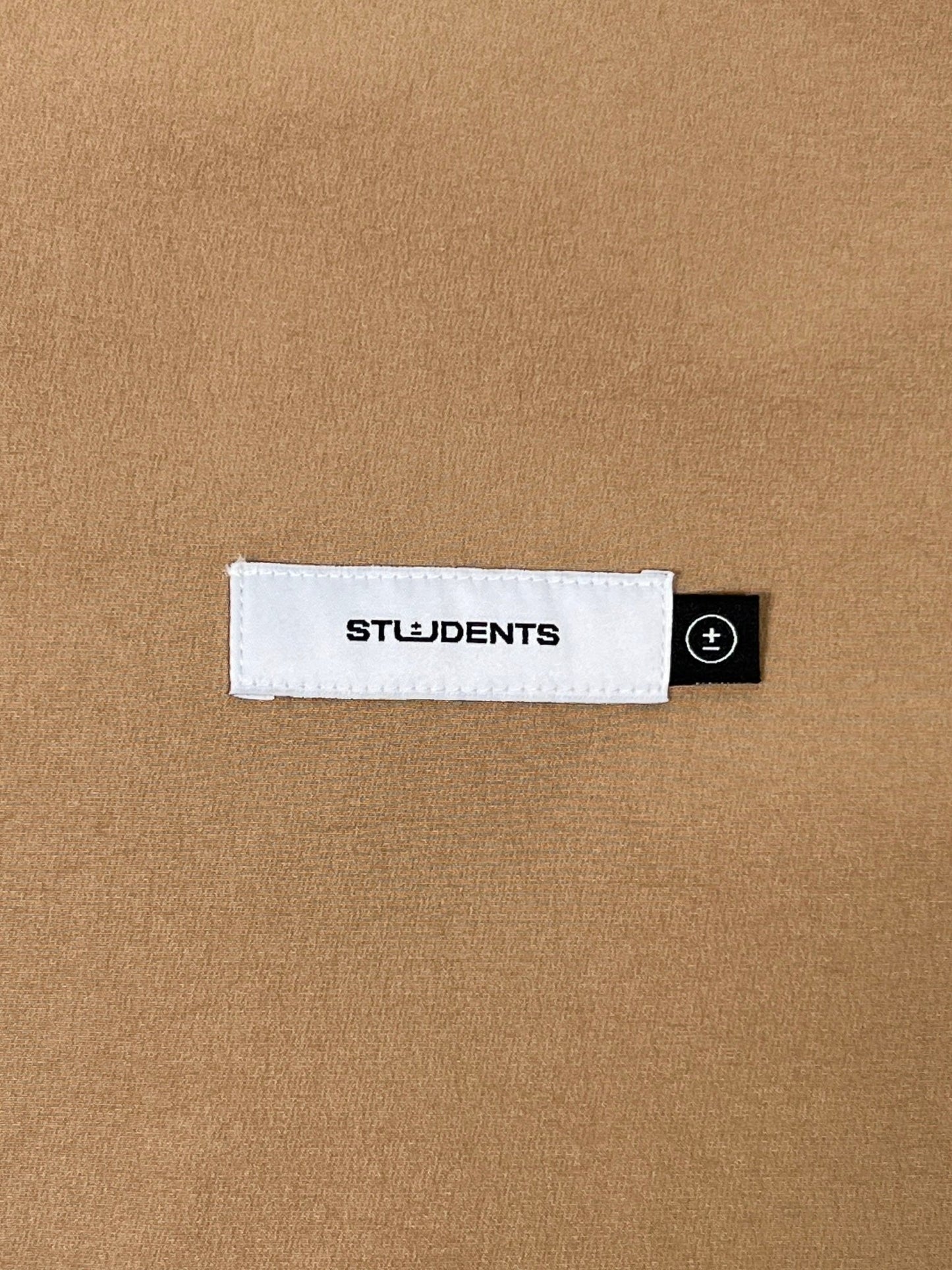 Close-up of the fabric from the Students Niall nylon popover jacket in tan, featuring a white rectangular patch with "STUDENTS" in bold black letters. It also includes a small black tag with a plus icon, and its design incorporates flexible stretch nylon for enhanced style and comfort.