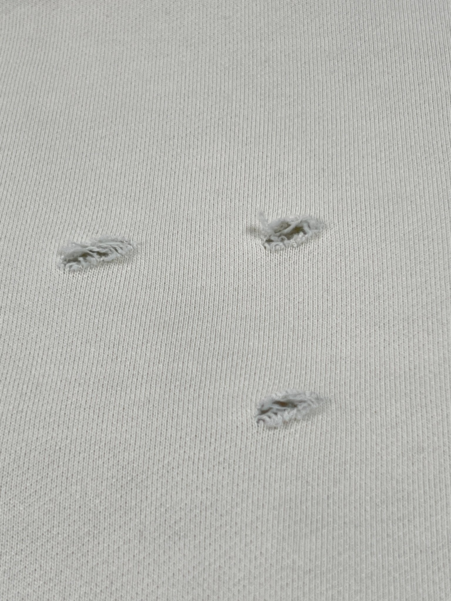 Close-up of the UNTITLED ARTWORKS Untitled Artworks Crewneck Short Sleeve Cream: light cream 100% cotton fabric with three frayed holes—ideal for a relaxed tee or short sleeve look.
