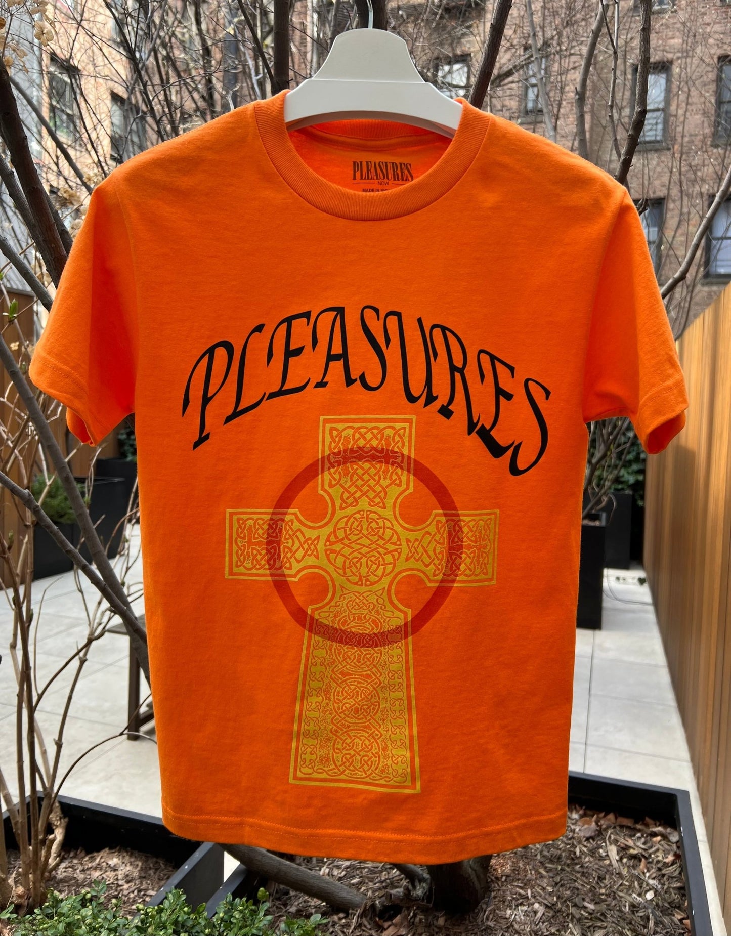 VEGAN T-SHIRT ORANGE by PLEASURES, with a large cross design in the center, displayed outdoors.