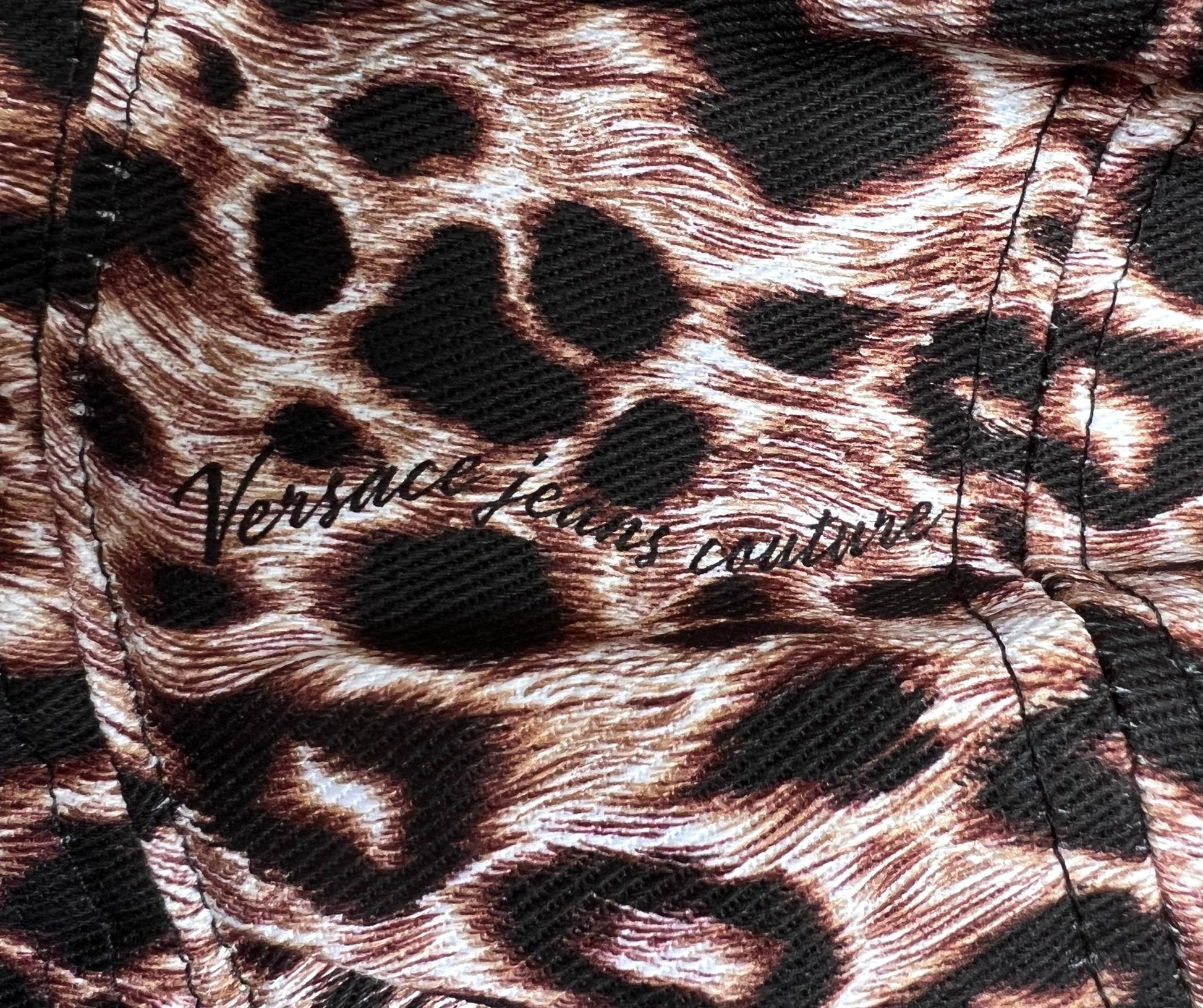 A close-up of the leopard print fabric captures the wild allure of the VERSACE E72GAZK08-EZG089-EY4V bucket hat, with "Versace Jeans Couture" elegantly printed in black over the design.