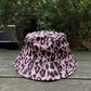 The VERSACE E72GAZK08-EZG089-EY4V bucket hat, featuring a chic leopard print, sits gracefully on a wooden table amid lush green foliage.