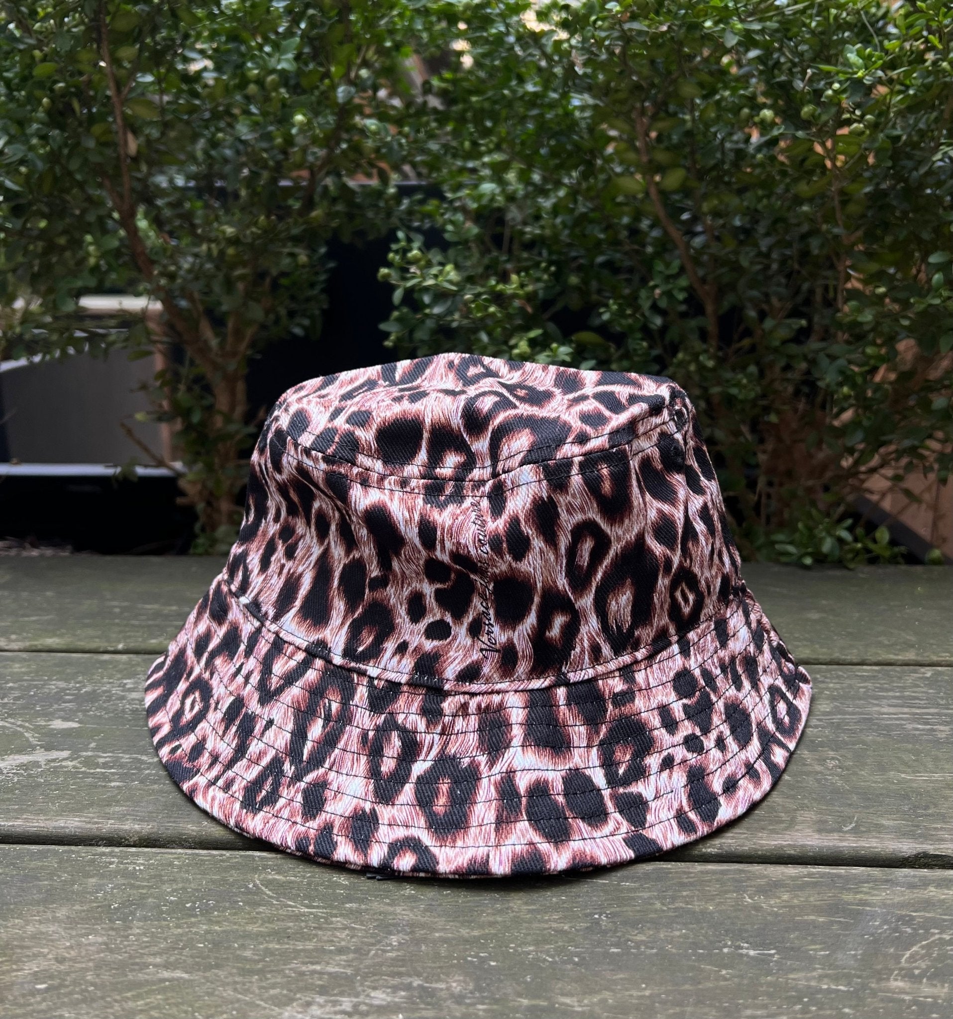 The VERSACE E72GAZK08-EZG089-EY4V bucket hat, featuring a chic leopard print, sits gracefully on a wooden table amid lush green foliage.