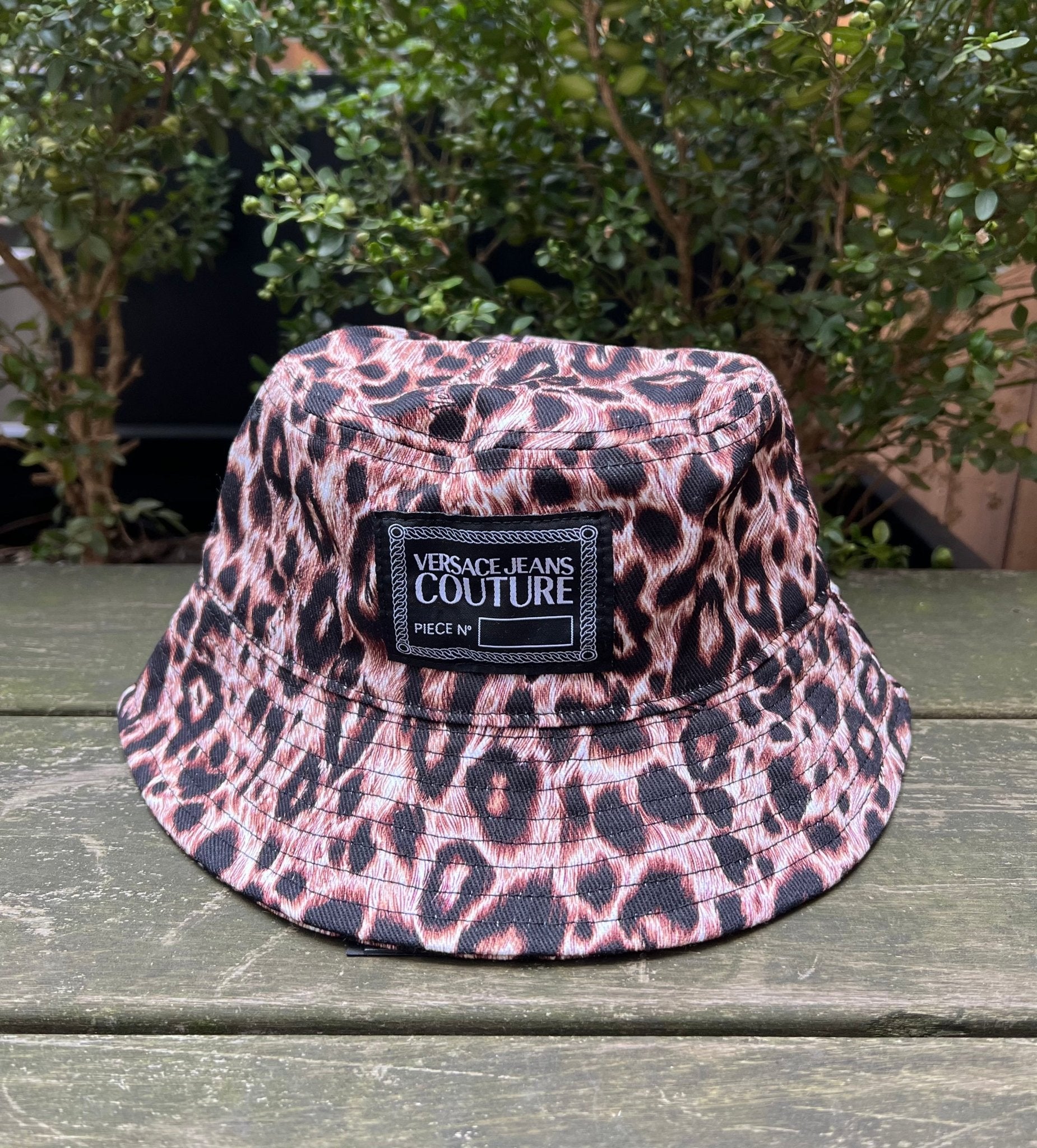 The VERSACE E72GAZK08-EZG089-EY4V bucket hat rests on a wooden surface, surrounded by lush greenery.