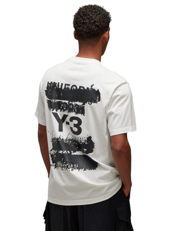 A person stands with their back turned, showcasing the Y-3 JM7815 U GFX SS Tee Cwhite by ADIDAS x Y-3, made from soft cotton fabric and adorned with a black abstract design and text on the back.