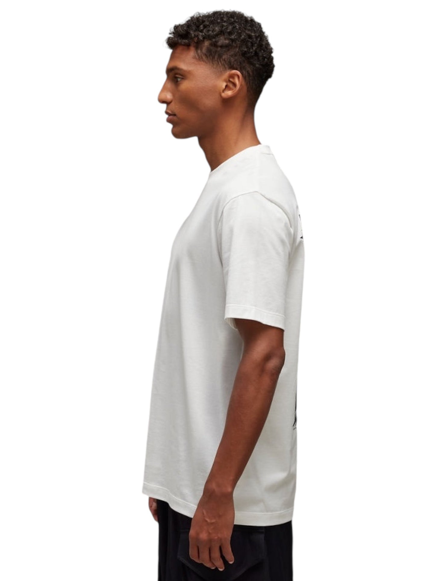 An individual stands in profile wearing the Y-3 JM7815 U GFX SS Tee Cwhite by ADIDAS x Y-3, crafted from soft cotton fabric, paired with black pants, set against a plain background.