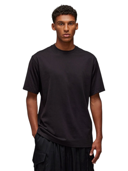 A person wearing the ADIDAS x Y-3 JN4879 U GFX SS Tee Black, crafted from soft cotton fabric, along with matching black pants, stands elegantly against a white backdrop, exuding the minimalist sophistication often seen in Y-3 designs.