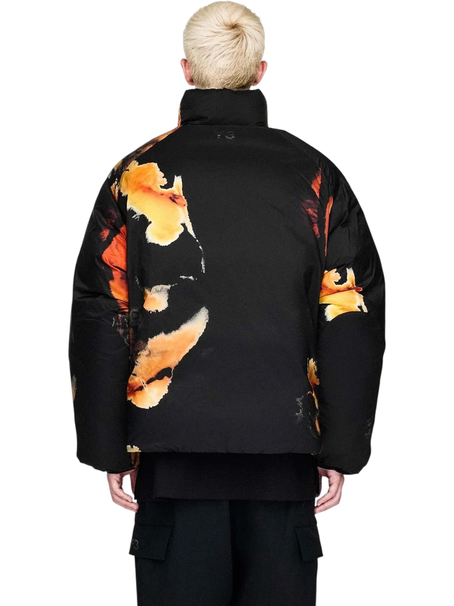 A person with short blond hair is seen from behind wearing the ADIDAS x Y-3 KA2364 M AOP Puff JKT Multco/Black, a puffer jacket with PrimaLoft insulation, black polyamide fabric, and bold orange-yellow abstract patterns.