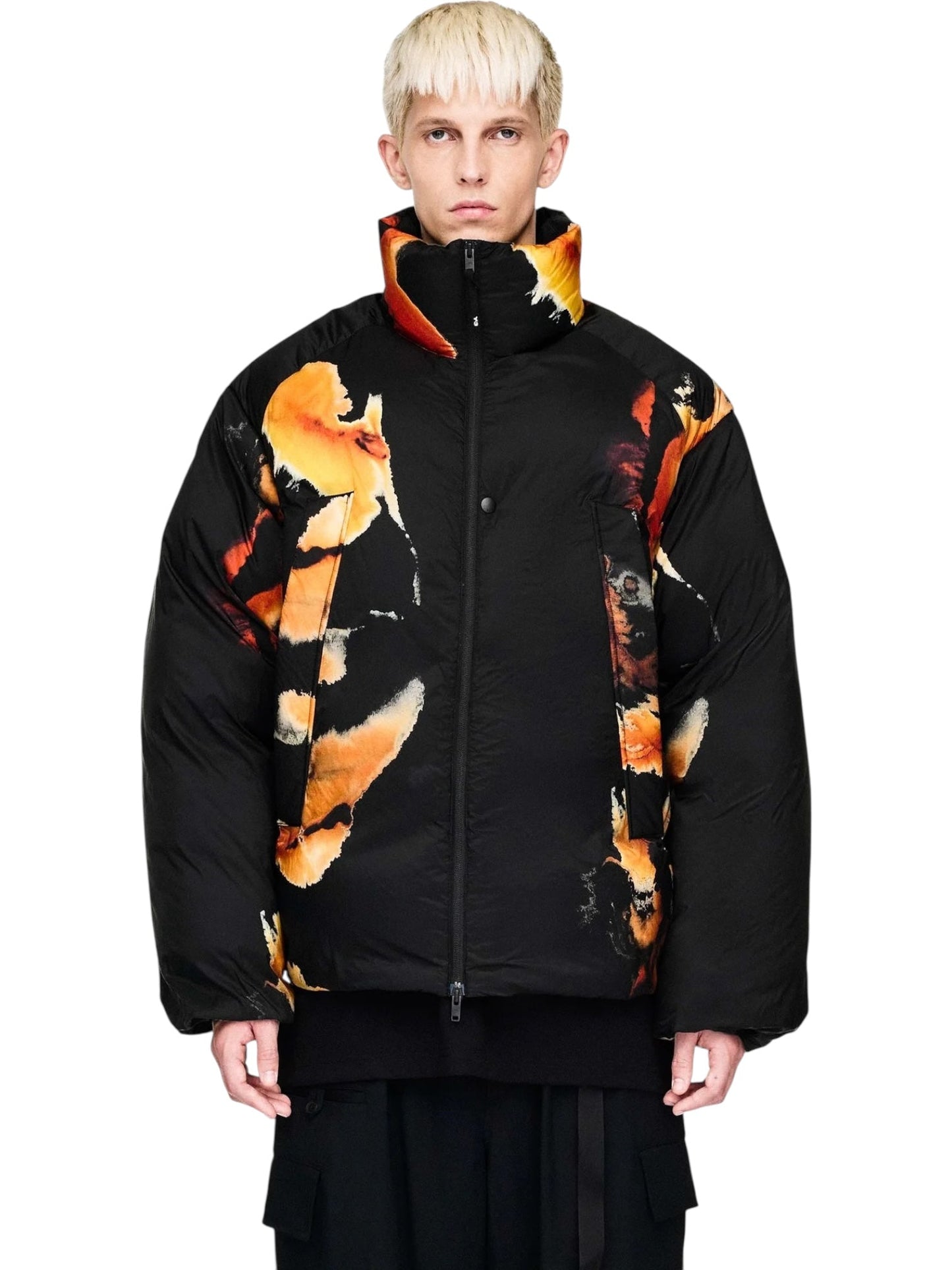 A person with short blond hair wears the ADIDAS x Y-3 KA2364 M AOP Puff JKT in Multco/Black, a black polyamide jacket with a bold orange and yellow abstract floral pattern, standing against a plain white background.