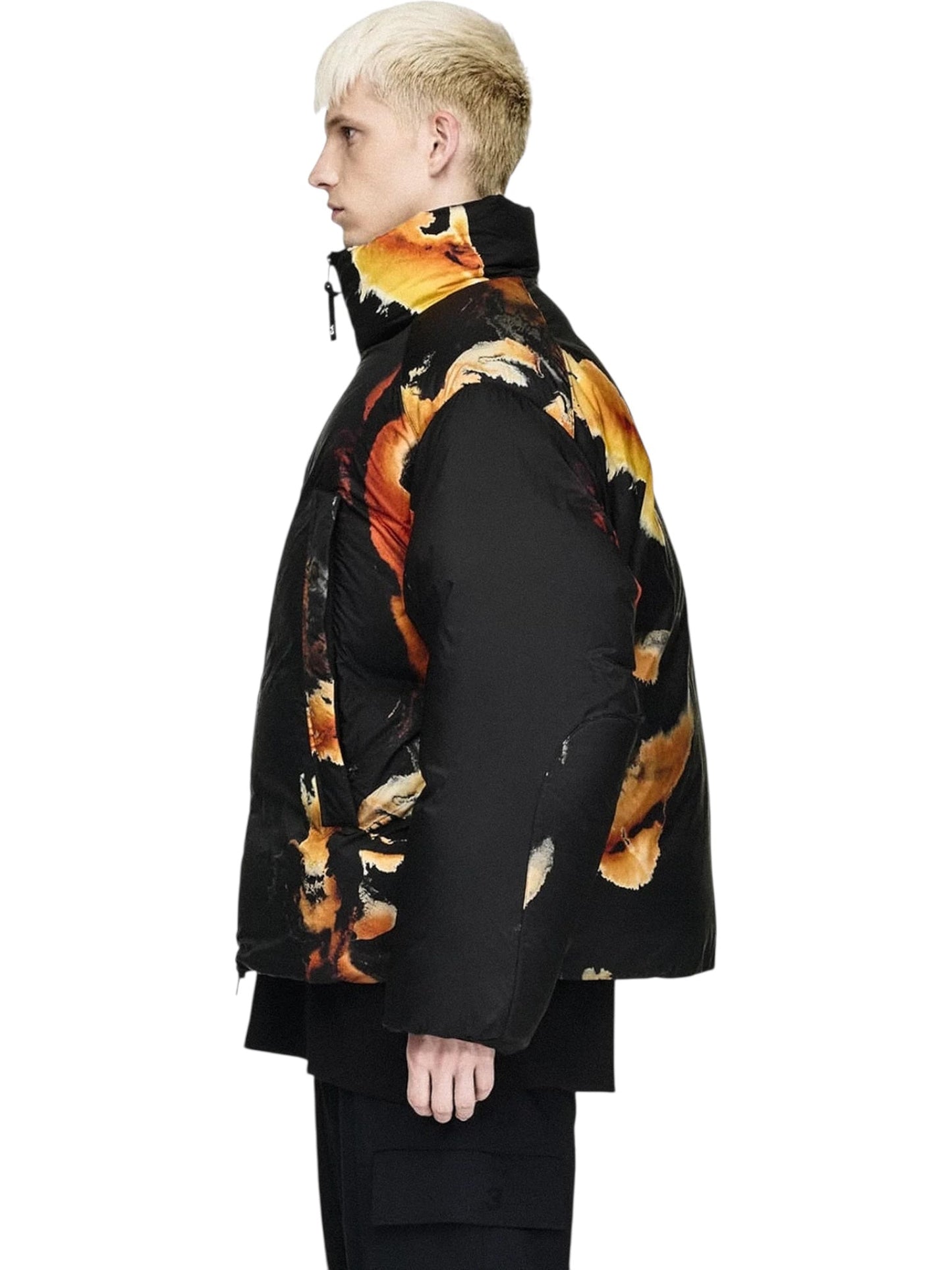 A person with short blond hair stands in profile, wearing ADIDAS x Y-3’s Y-3 KA2364 M AOP Puff JKT Multco/Black—a black puffer jacket with an abstract orange and yellow pattern—and black pants.