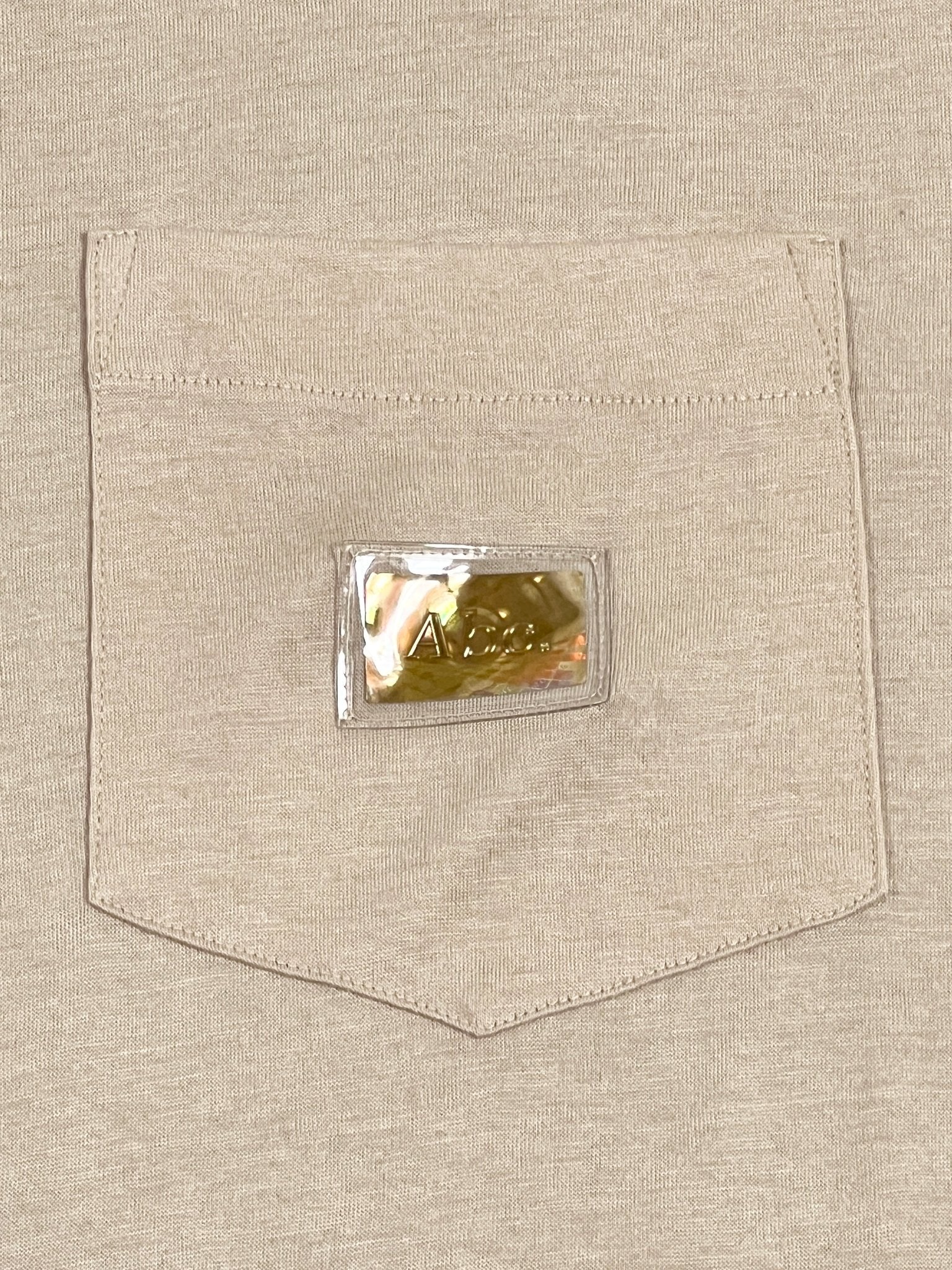 A beige Advisory Board Crystals S/S Pocket Tee Feldspar Ecru from ADVISORY BOARD CRYSTALS featuring a cotton shirt pocket with a small, transparent plastic pouch holding a gold item inside.