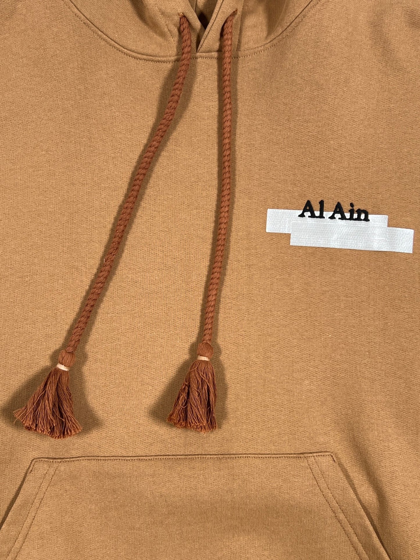 Close-up of a brown AL AIN AHOX S102 PALMIER CHAMEAU hoodie with drawstrings and a label reading "al ain".