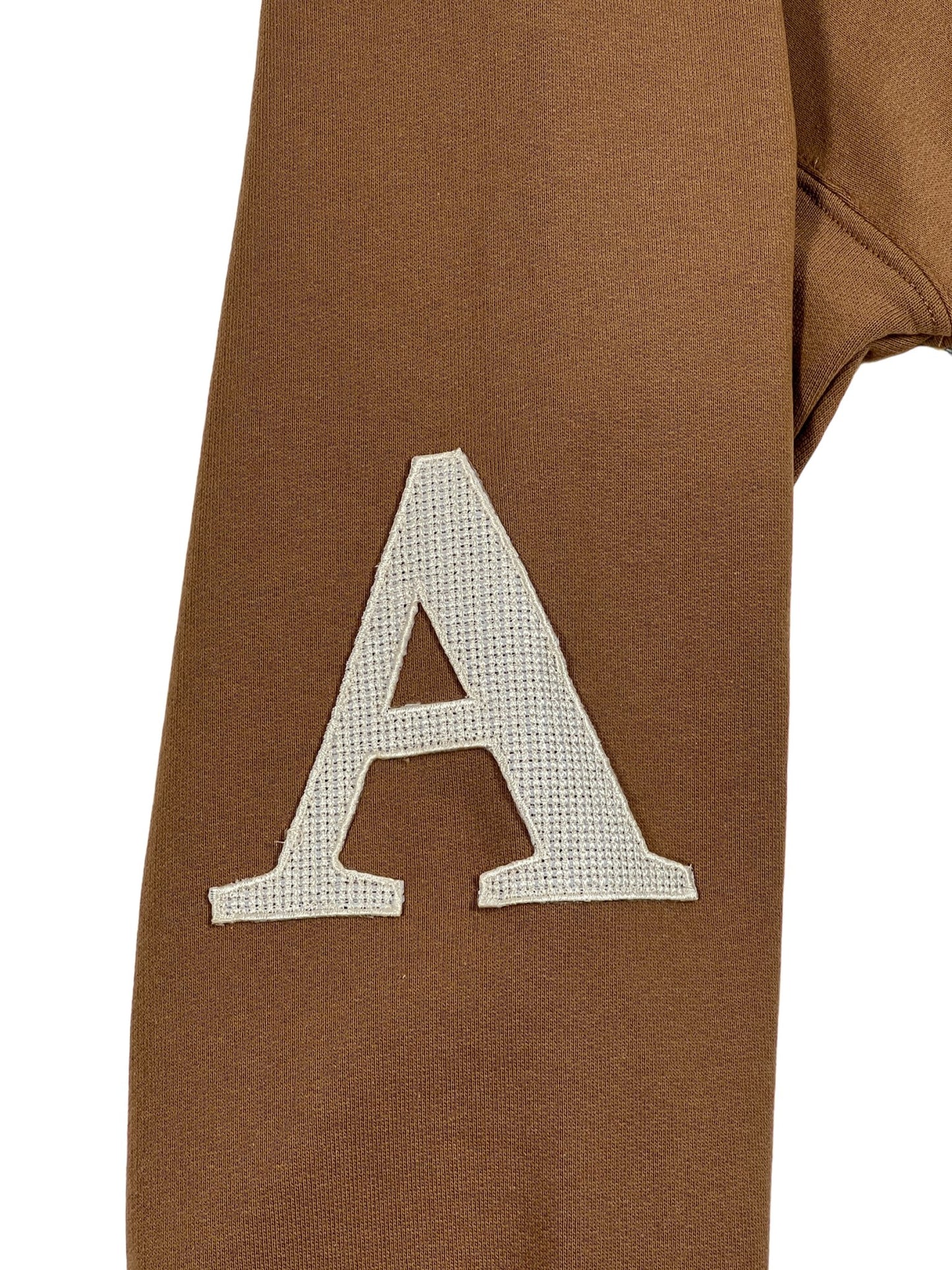 Close-up of a white letter 'a' patch sewn onto a brown AL AIN AHOX S102 PALMIER CHAMEAU hoodie.