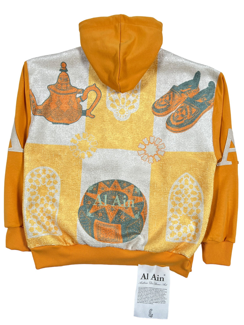 Gold and ocre yellow AL AIN hoodie with traditional Arab motifs, including a teapot and slippers, labeled "AL AIN".