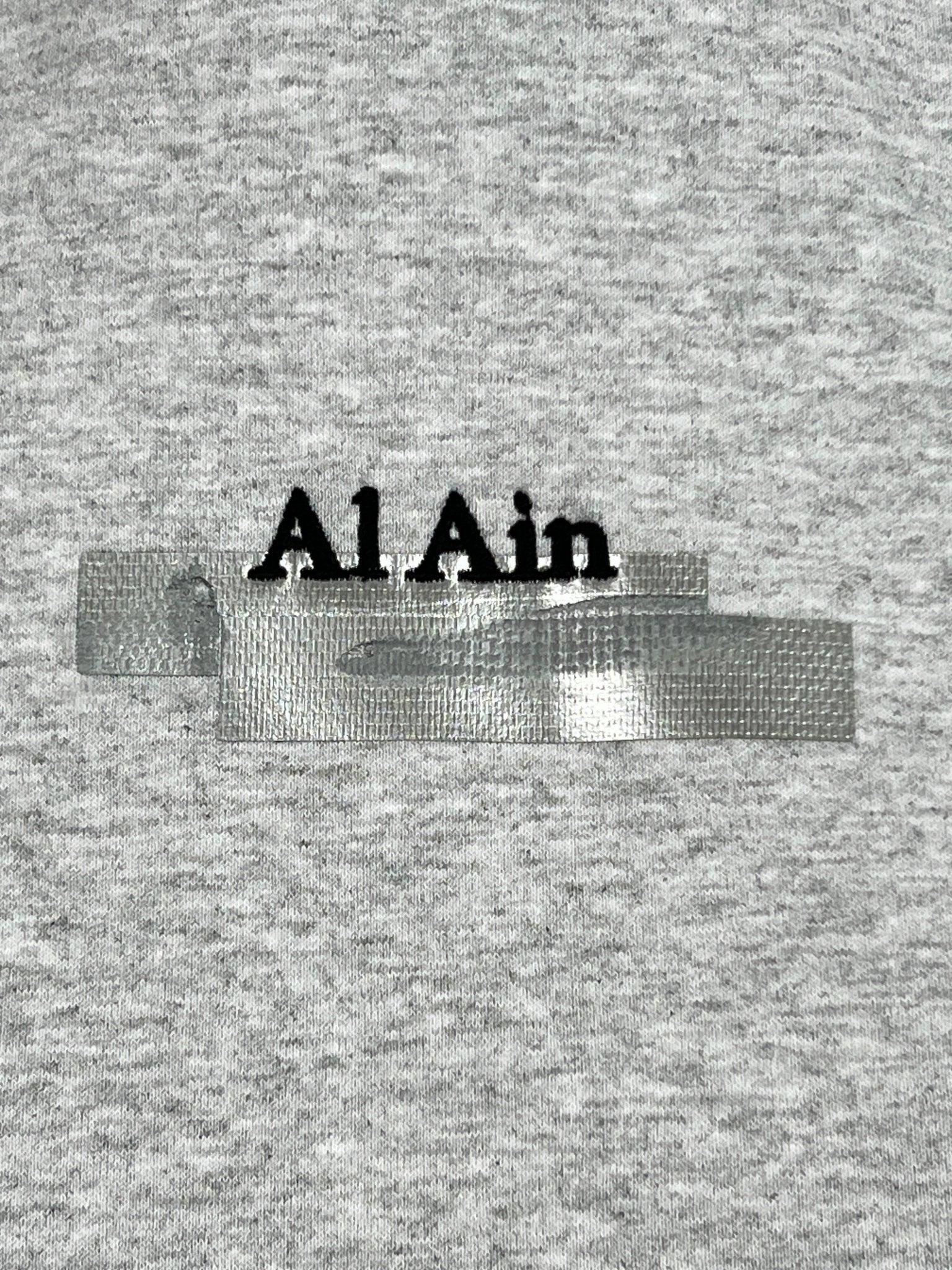 Close-up of a gray AL AIN hoodie with the words "AL AIN AHOX S150 YACHT LIMITED" embroidered in black. The embroidery is partially covered by two pieces of silver tape.