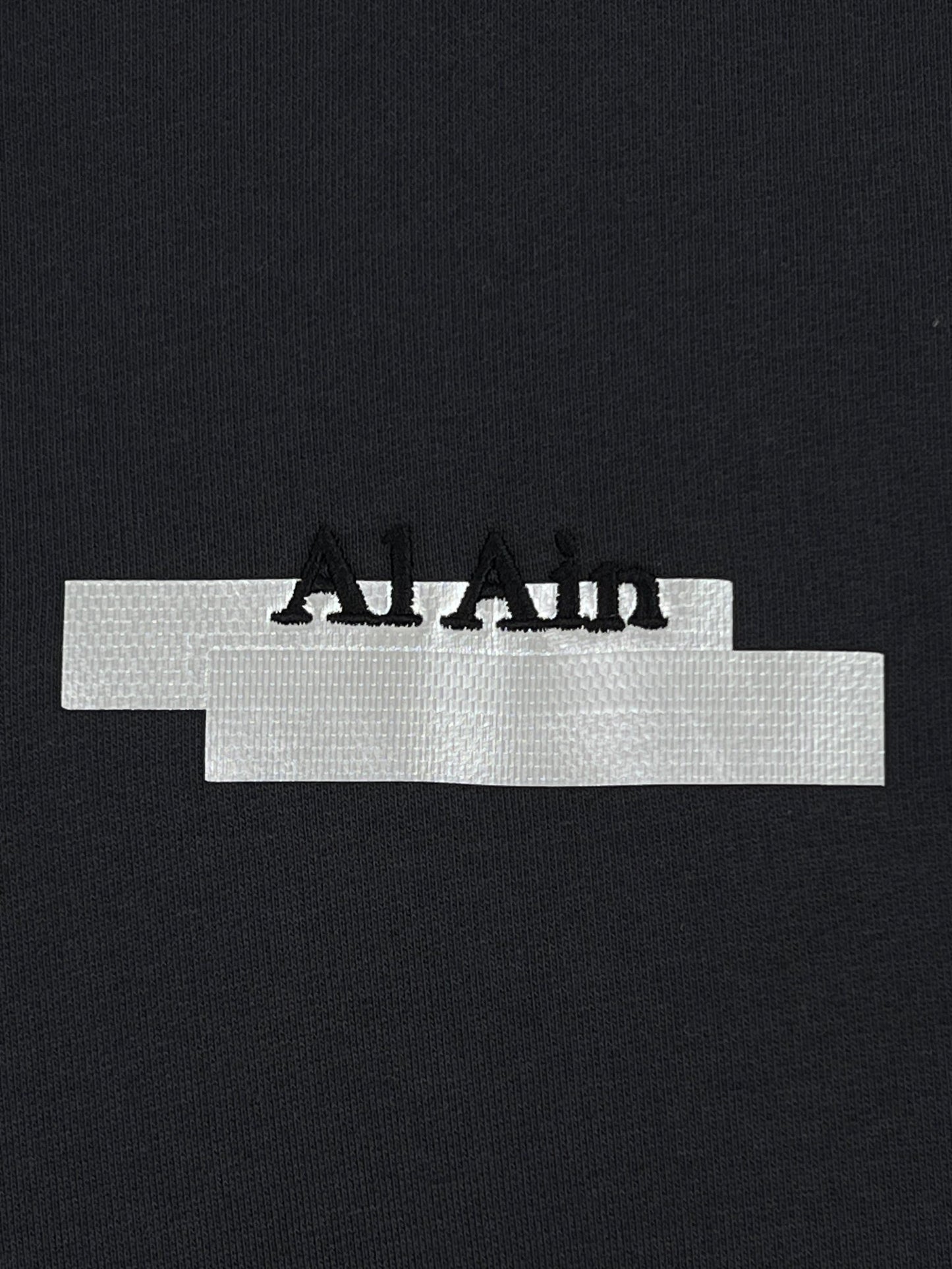 Close-up of a black fabric texture with a white label bearing the text "AL AIN AHZX S103 SECRET NOIR" in black letters.
