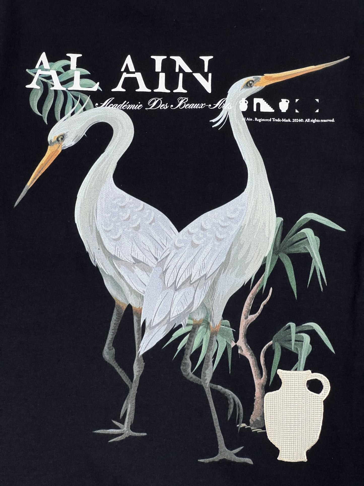 Graphic print of two herons on a black AL AIN AMHX S120 JABALIA NOIR graphic t-shirt with embroidered brand name, decorative text, and foliage.