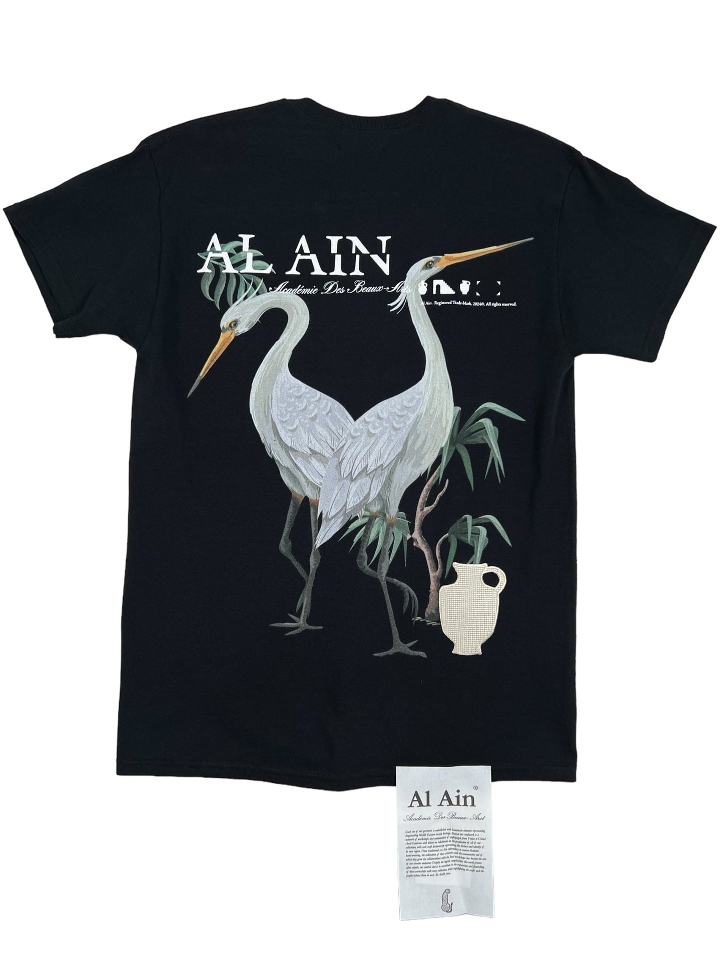 Black AL AIN t-shirt with an embroidered AL AIN AMHX S120 JABALIA NOIR graphic design featuring a white heron, displayed with a clothing tag visible at the bottom.