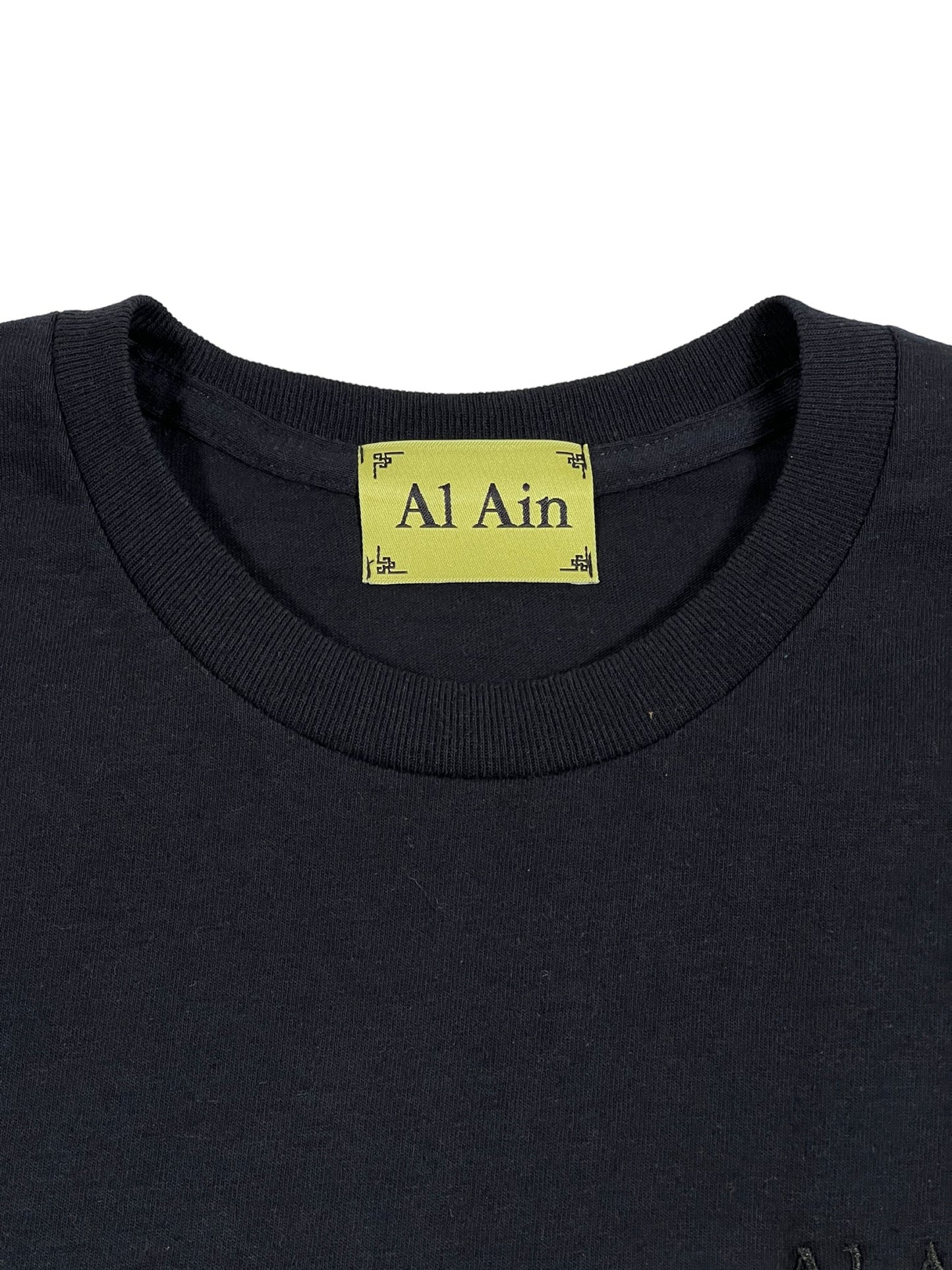 Close-up of a black AL AIN AMHX S120 JABALIA NOIR graphic t-shirt's neckline with an embroidered AL AIN brand name label.