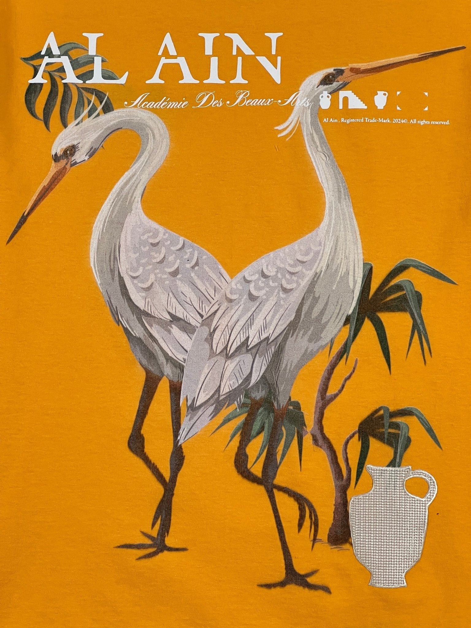 Two white herons stand among green plants, with bold "AL AIN AMHX S120 Jabalia Ocre Yellow" branding and a white vase on an orange background—perfect for a standout AL AIN graphic tee.