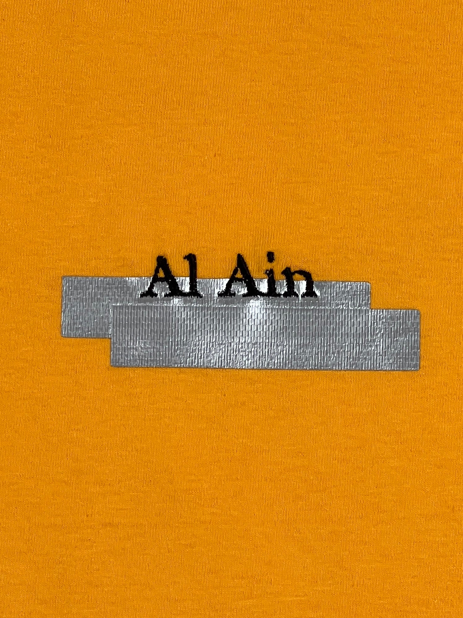 The AL AIN "Al Ain AMHX S120 Jabalia Ocre Yellow" features the text "Al Ain" partially covered by two overlapping silver duct tape strips on an ochre yellow background, creating a bold, graphic T-shirt-inspired detail.