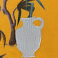 The Al Ain AMHX S120 Jabalia Ocre Yellow by AL AIN features a bold, artistic design with a white textured jug on a mustard yellow background, accented by green leaves and brown branches in the upper left corner.