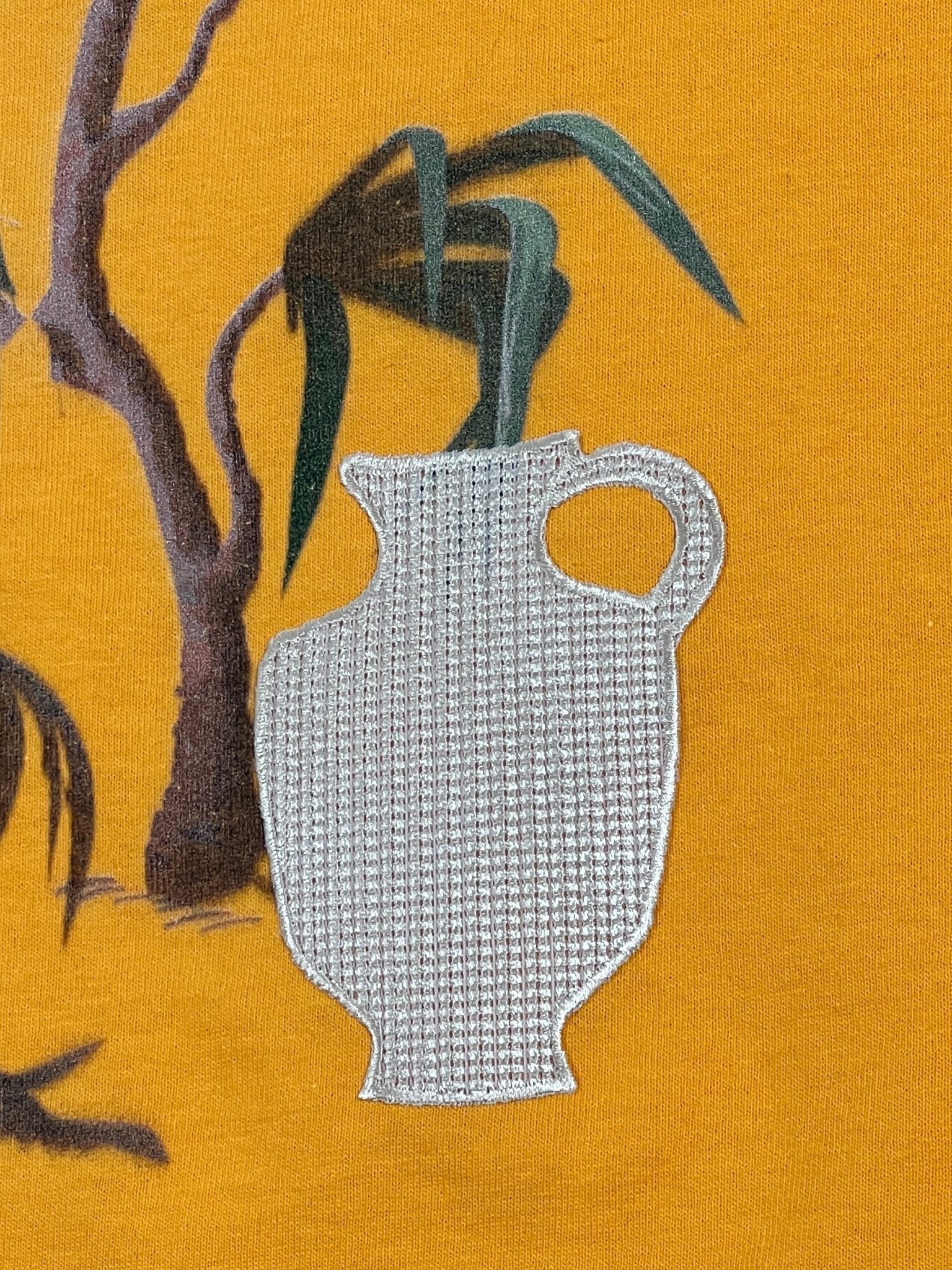 The Al Ain AMHX S120 Jabalia Ocre Yellow by AL AIN features a bold, artistic design with a white textured jug on a mustard yellow background, accented by green leaves and brown branches in the upper left corner.