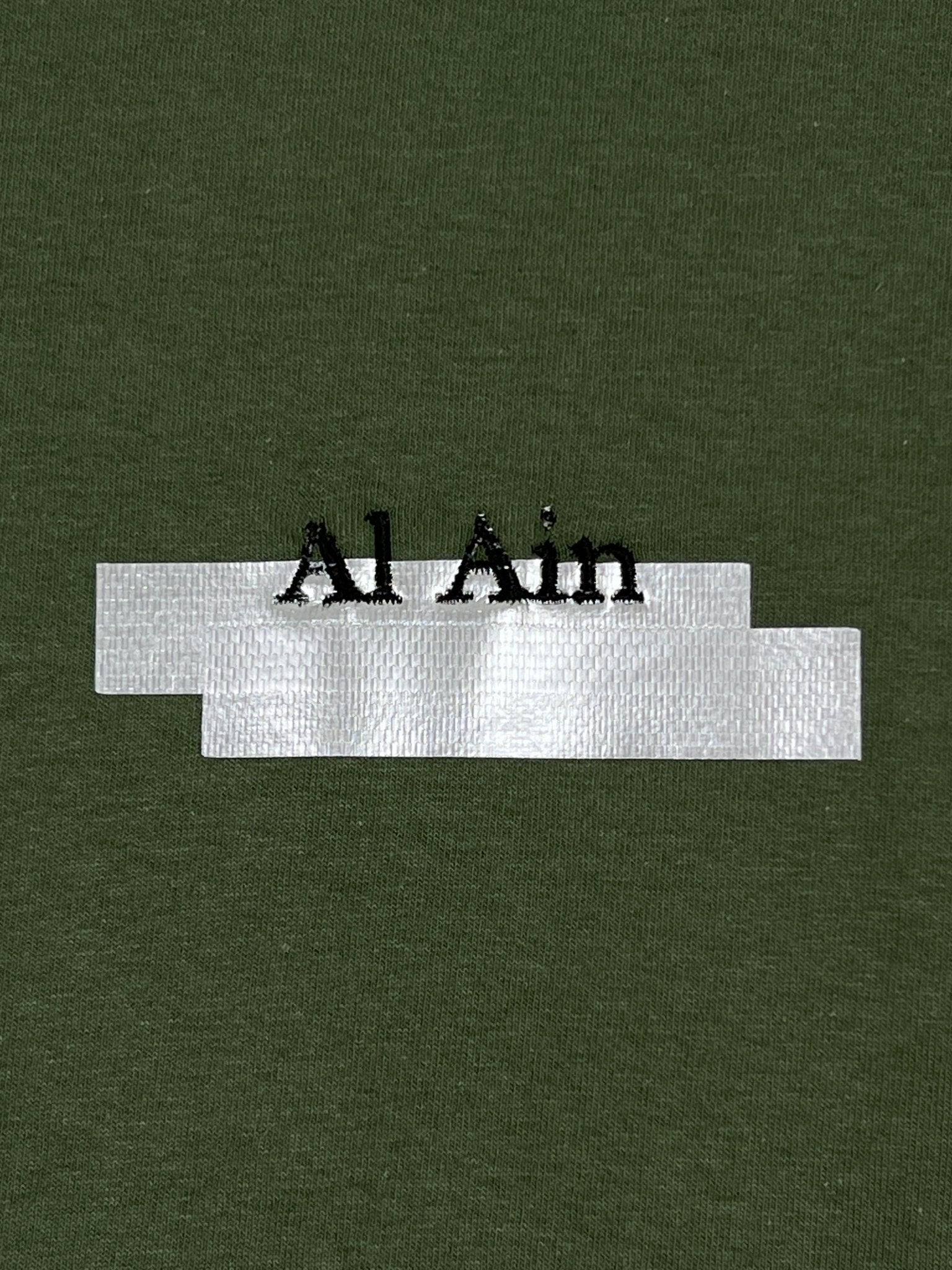 On an olive green background, a white band showcases the bold black lettering "Al Ain," representing the Al Ain AMHX S121 Peacock Militaire by brand AL AIN.