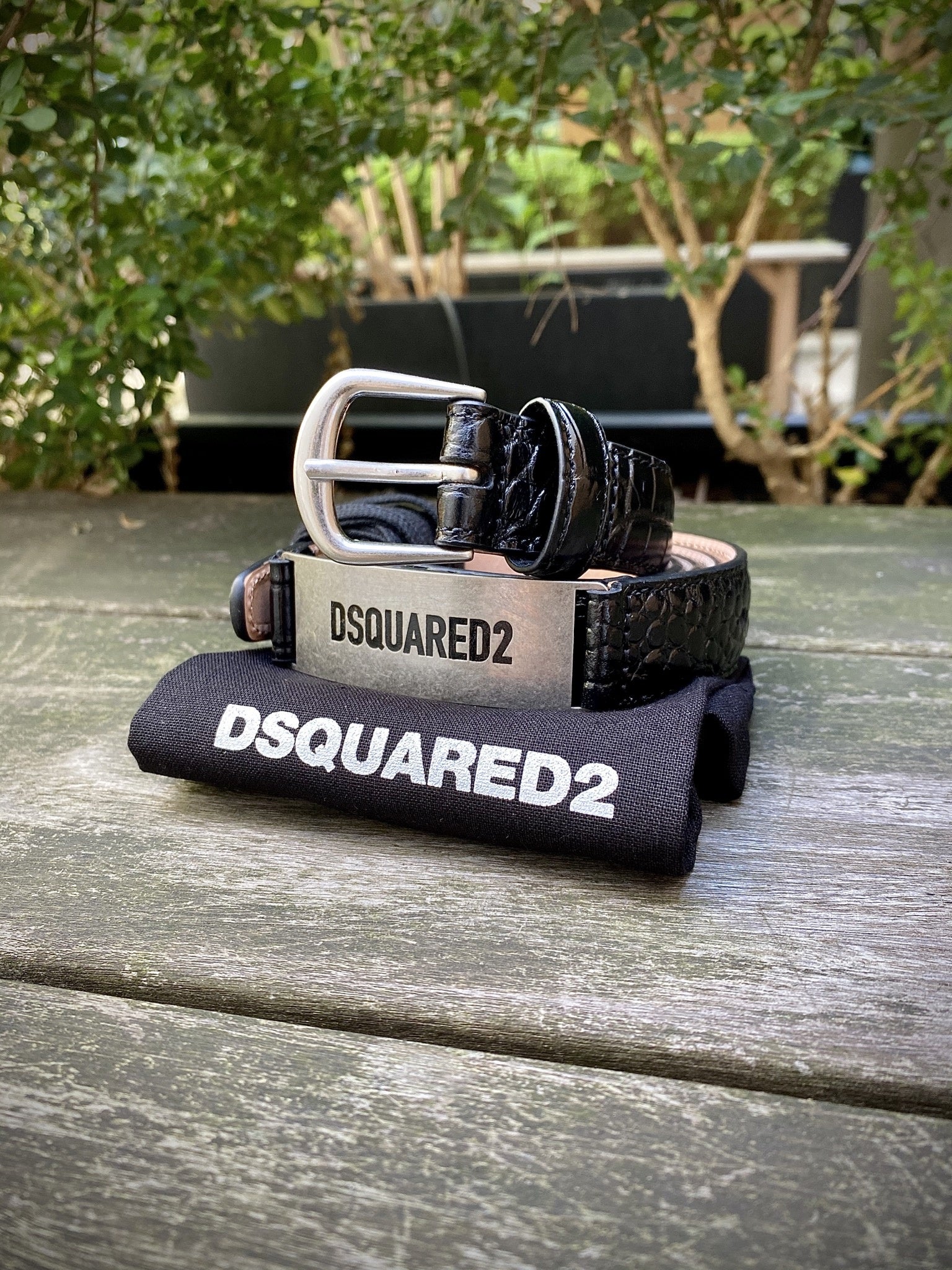 The Dsquared2 BEM0370 Belt-Vitello Stampa Cocco Nero by DSQUARED2, showcasing a refined silver buckle and elegant crocodile print, is beautifully arranged on a coordinating black cloth atop a wooden surface, amid lush greenery. Crafted in Italy, this piece epitomizes sophistication and expert artistry.