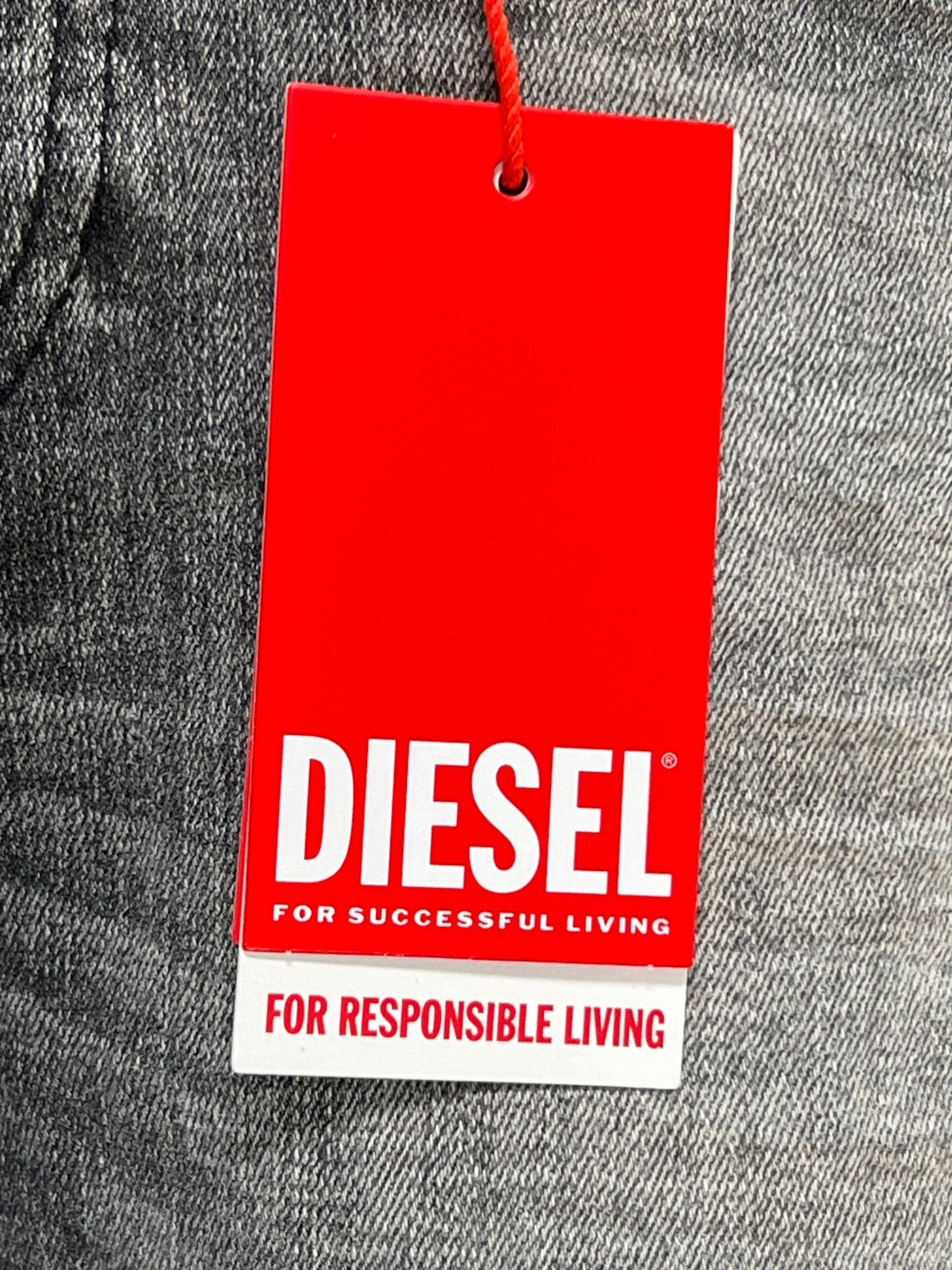 The Diesel 1979 Sleenker 9H74 skinny jeans, made from organic cotton stretch denim, feature a red tag with "DIESEL" and the phrases "For Successful Living" and "For Responsible Living," set against a gray fabric backdrop.