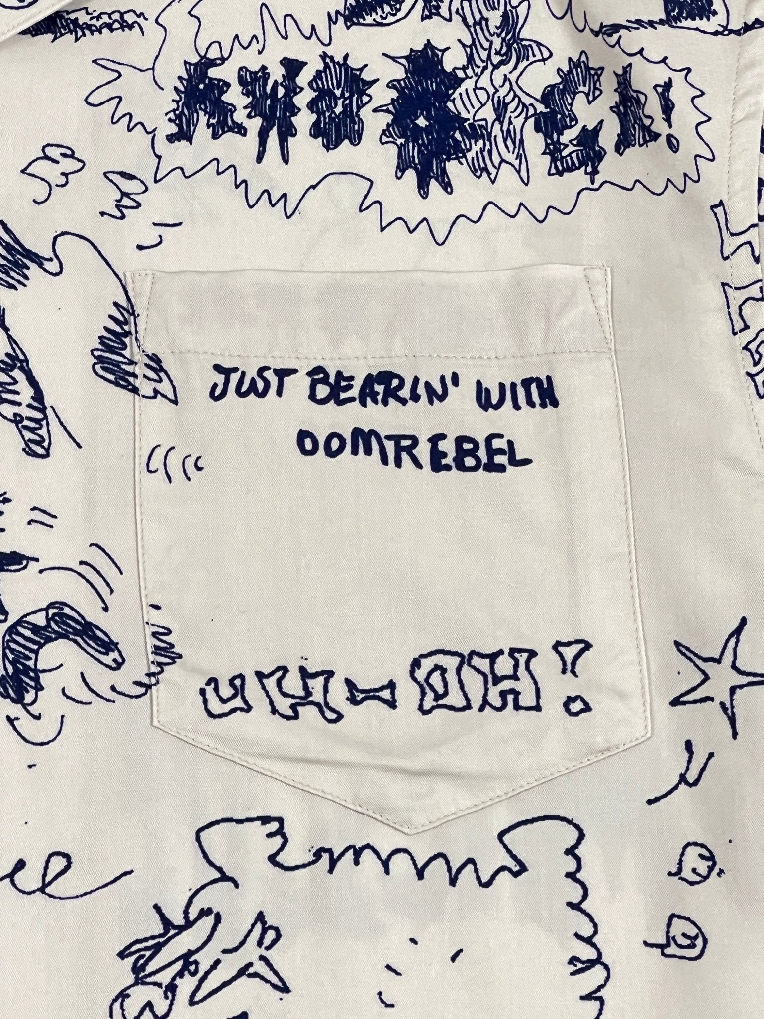 Crafted from white rayon and Tencel fabric adorned with blue doodles, the Domrebel Bearing Camp Collar Shirt in Beige by DOM REBEL features a short sleeve design and a stylish pocket with the text "JUST BEARIN' WITH OOMREBEL" and "UH-OH!" Its distinctive style makes it perfect for a casual day out.