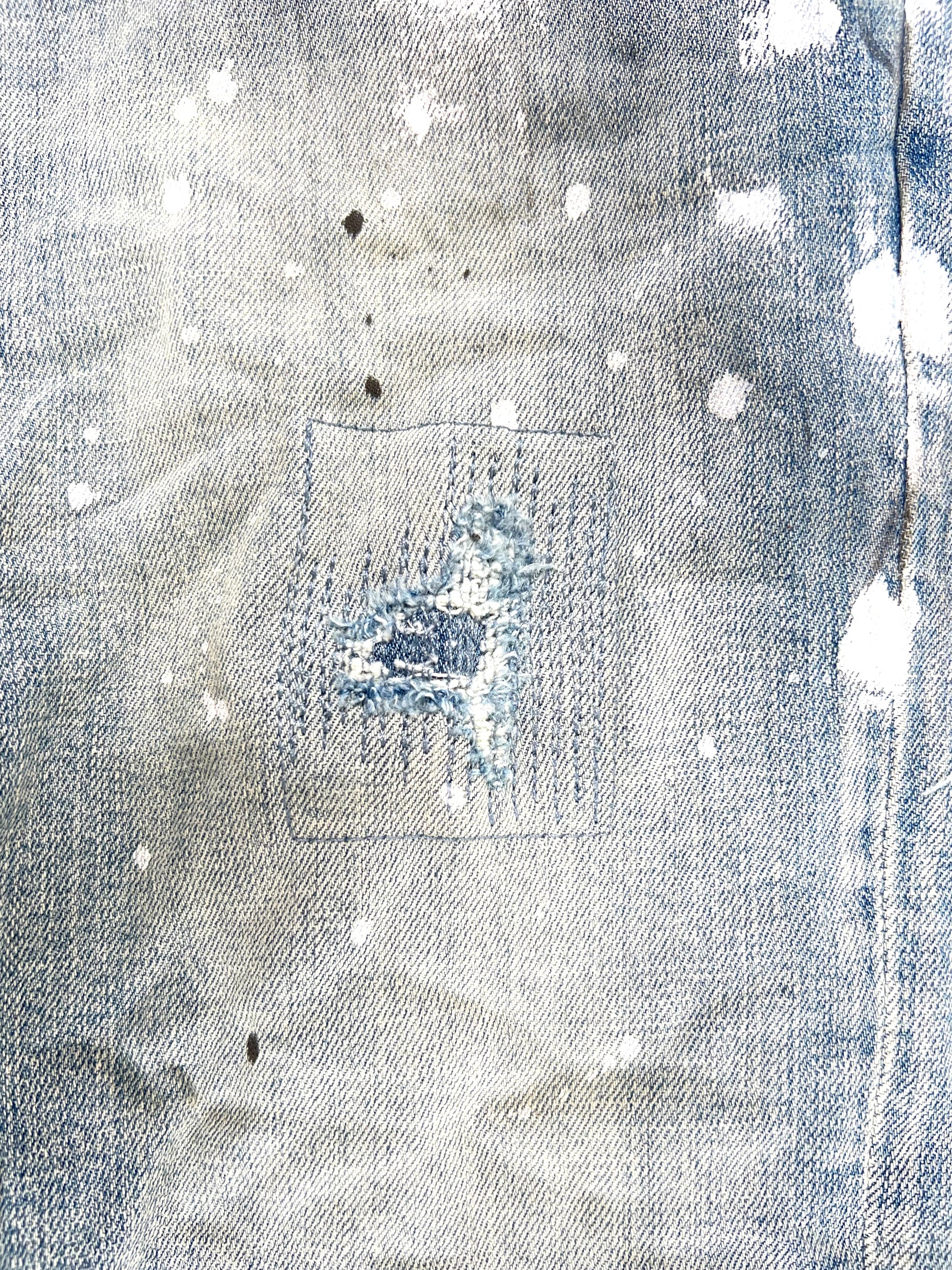 Close-up of the Purple Brand Jeans P001-LIA Light Indigo Paint Blowout from PURPLE BRAND, featuring paint splatters, a visible rectangular patch repair, and the edgy style of ripped jeans.