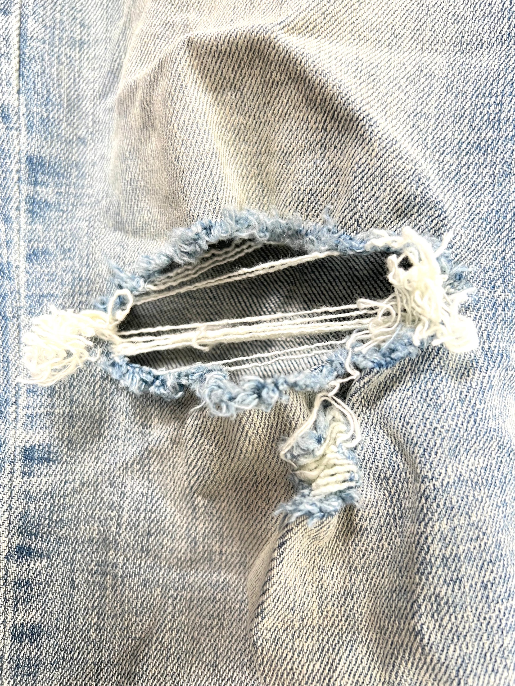 Close-up of a frayed hole in the Purple Brand Jeans P001-LIA Light Indigo Paint Blowout by PURPLE BRAND, with white threads exposed across the gap.