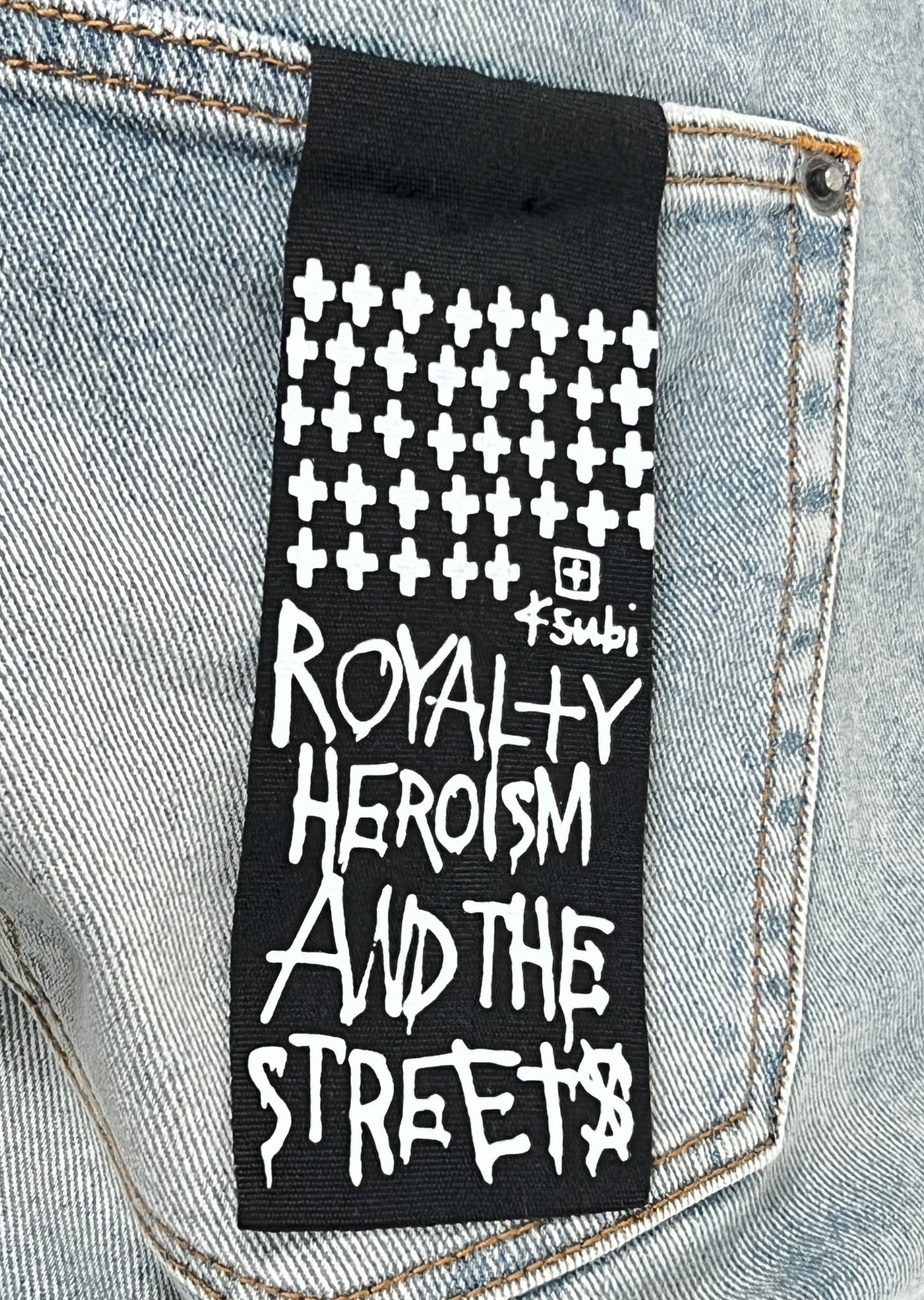 Black fabric label with white text reading "royalty heroism and the streets" sewn onto Ksubi Bronko Dynamite Metal Denim fabric.